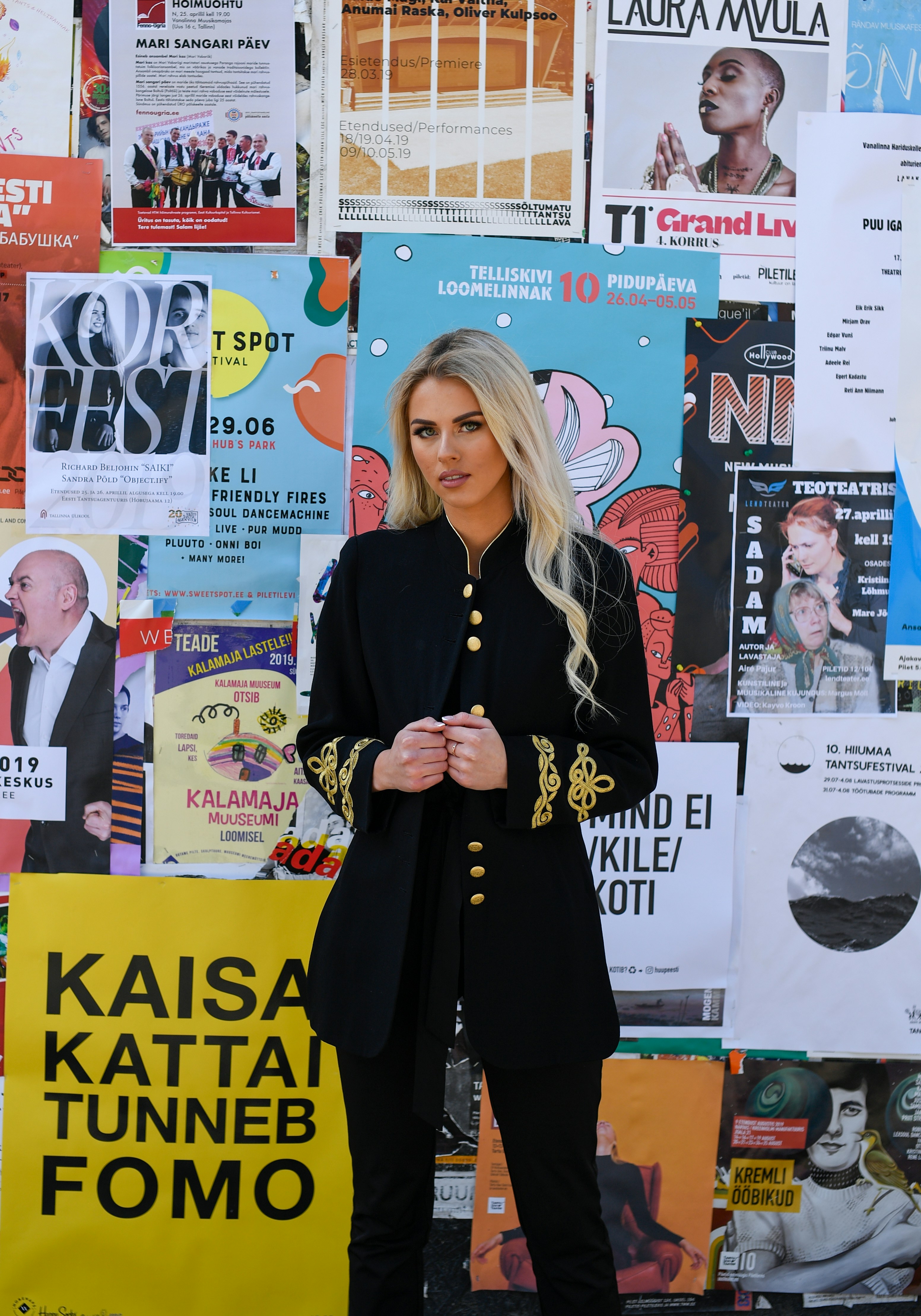 Une femme en veste noire ornée se tient face à des affiches colorées