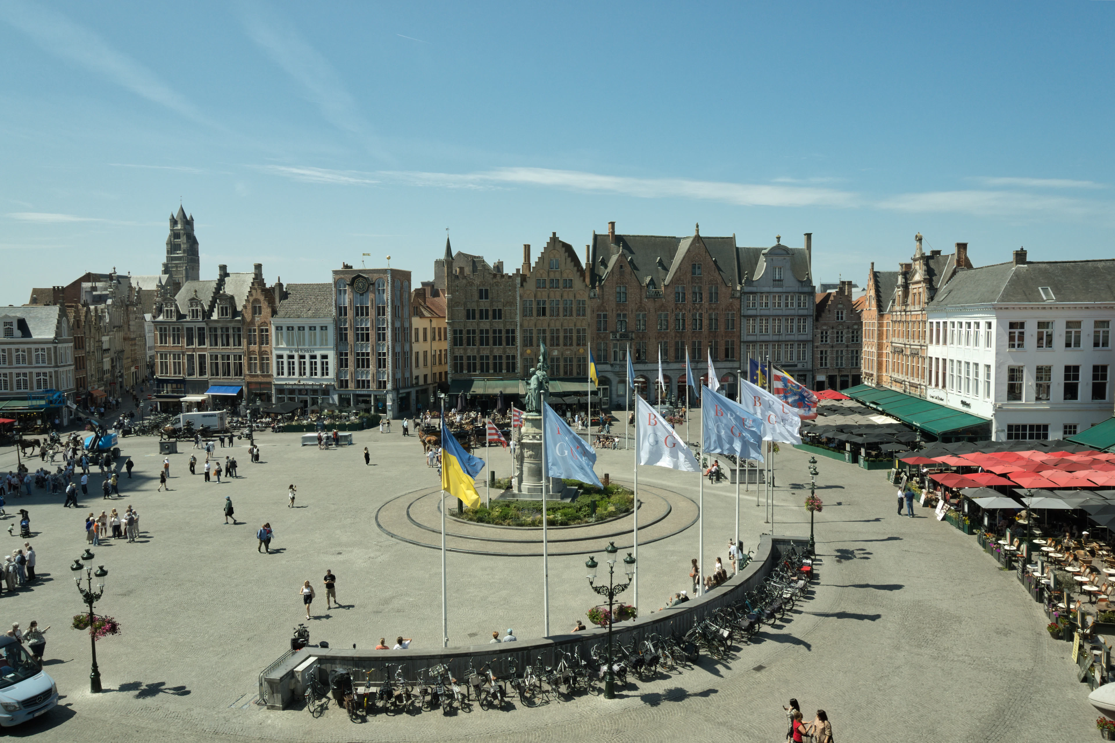 An elevated view over Bruges' Markt square with its colorful guild houses, flags and crowds on a sunny day.