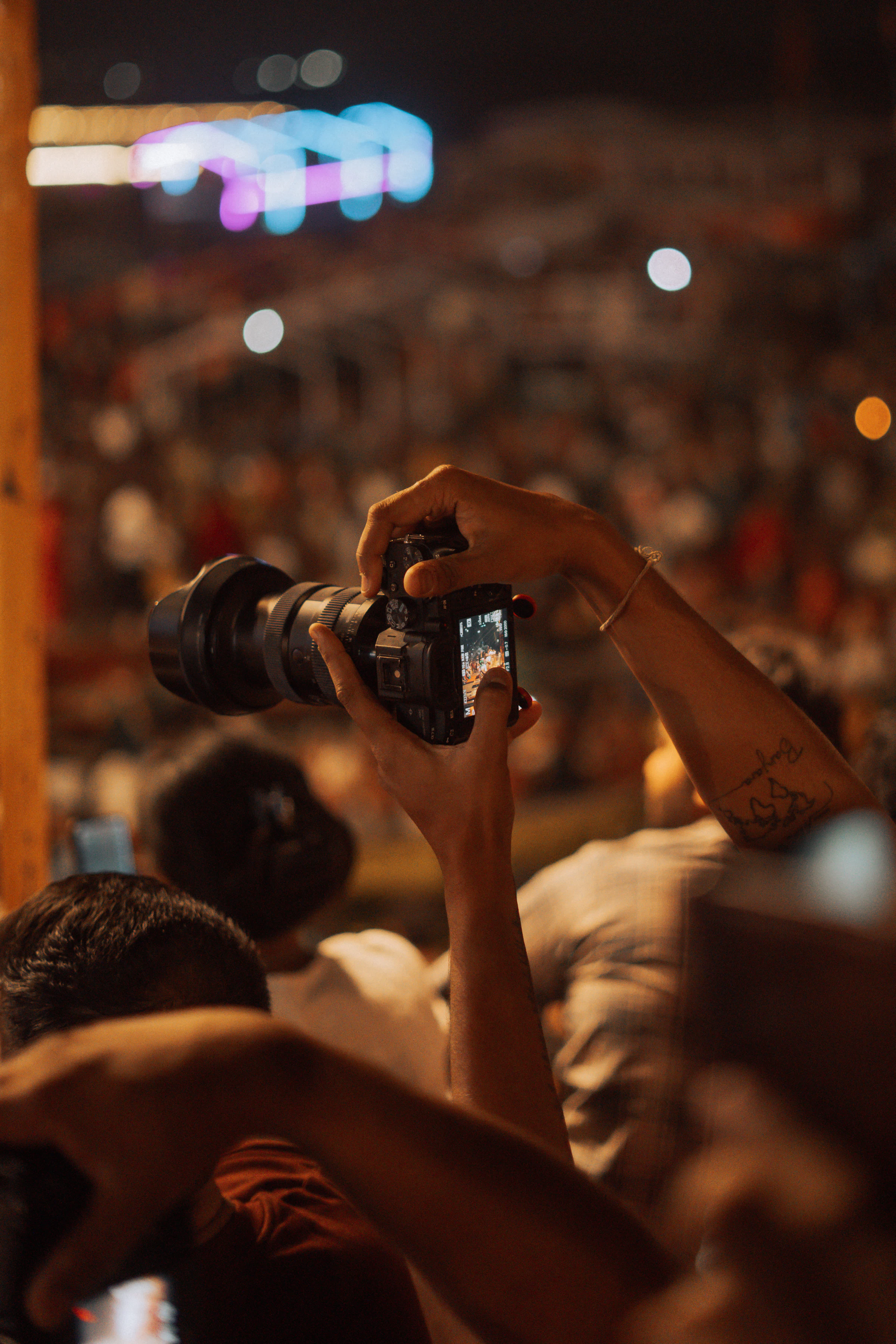 Hands raise a camera above a crowd at a night-time event in Varanasi, with bright lights behind
