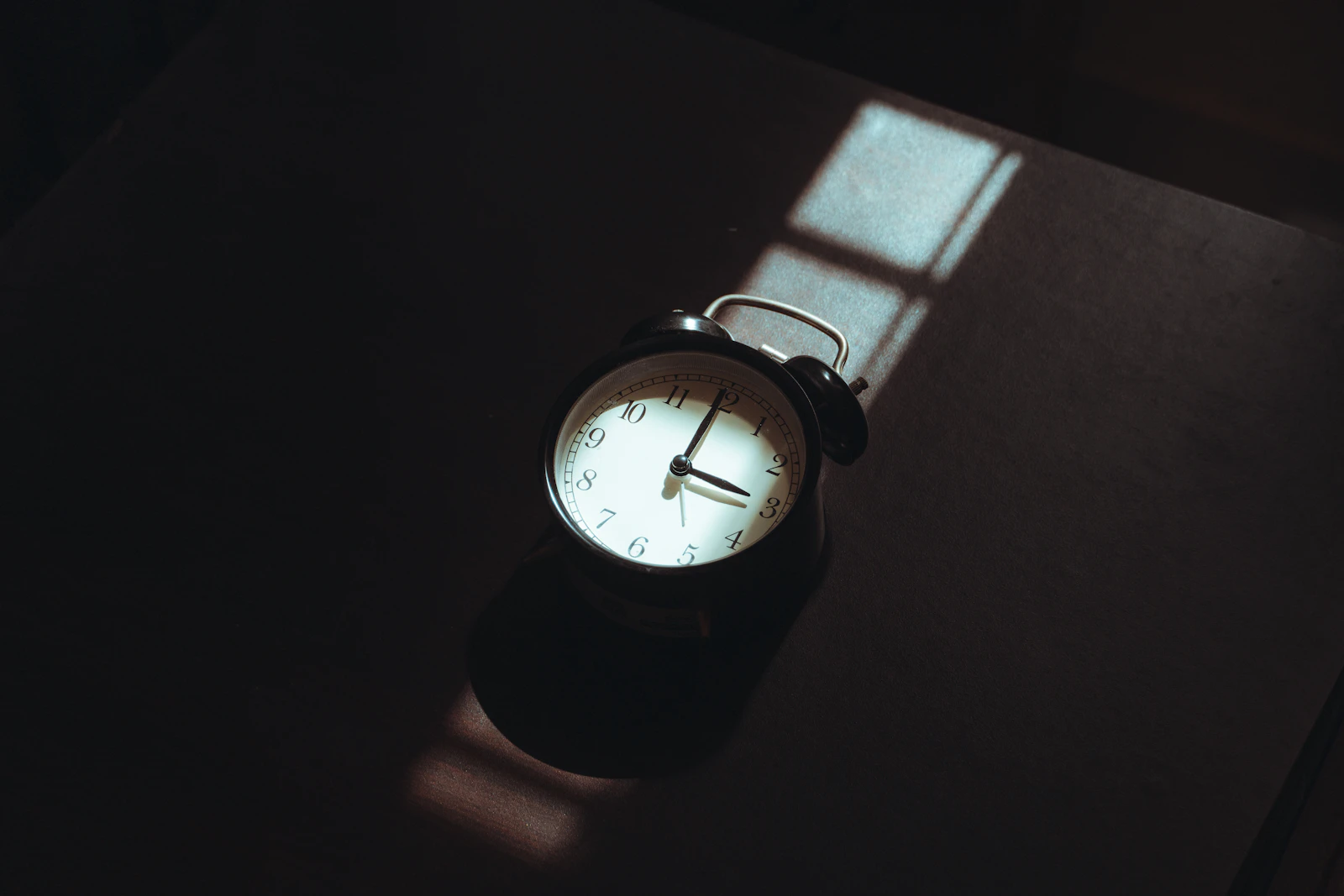 A small analogue alarm clock on a dark table, lit by a narrow shaft of window light