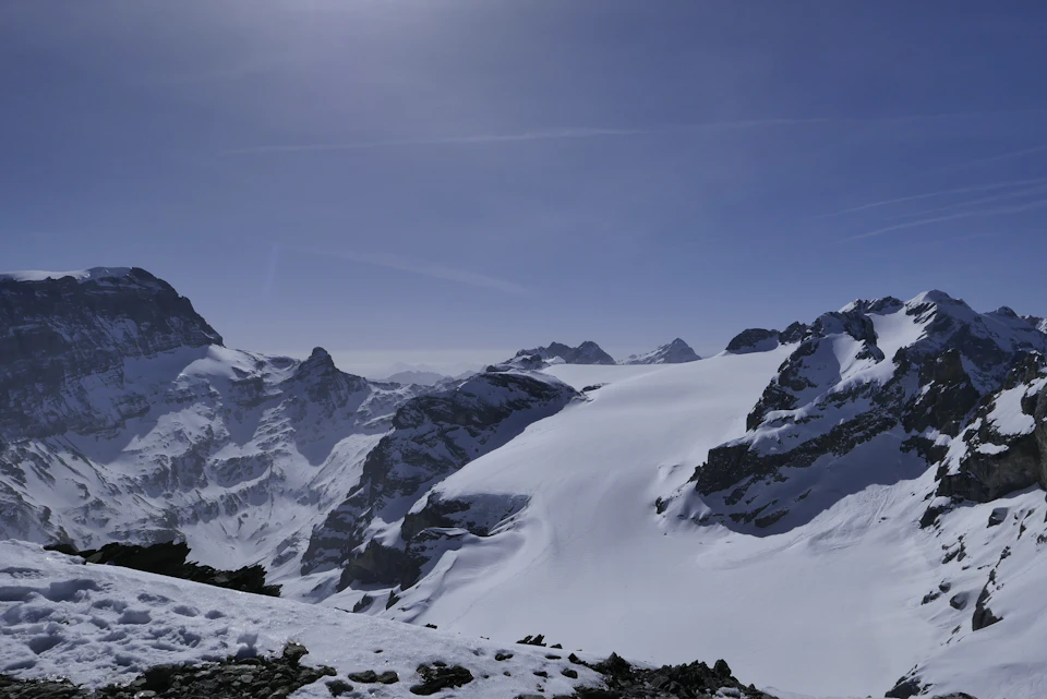 Snow-covered mountain range under clear blue sky with sun casting long shadows on white snow