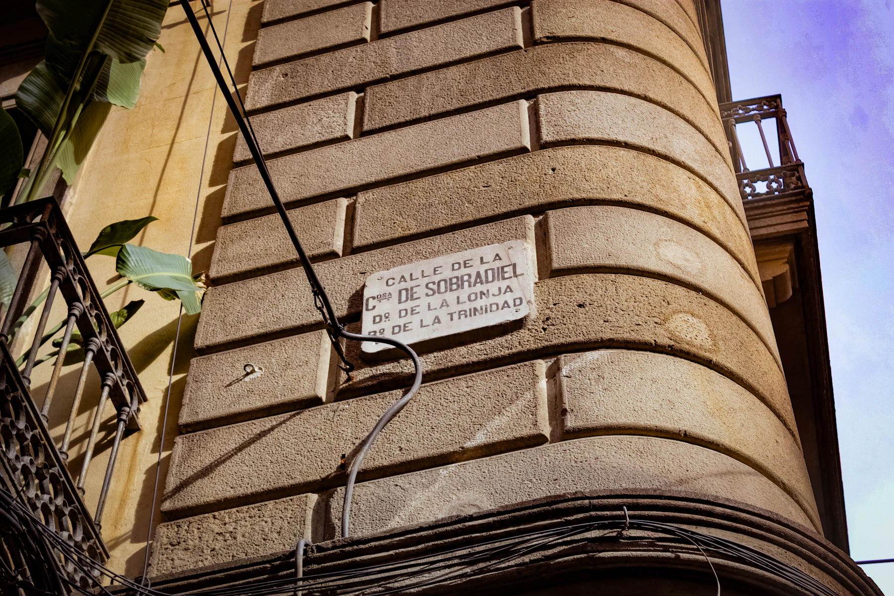 A rounded stone building corner with a white street plaque lettered in Spanish, a balcony and cables beside it
