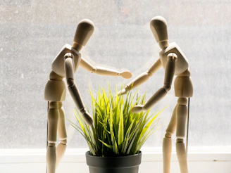 Two wooden mannequins reaching towards a potted plant.