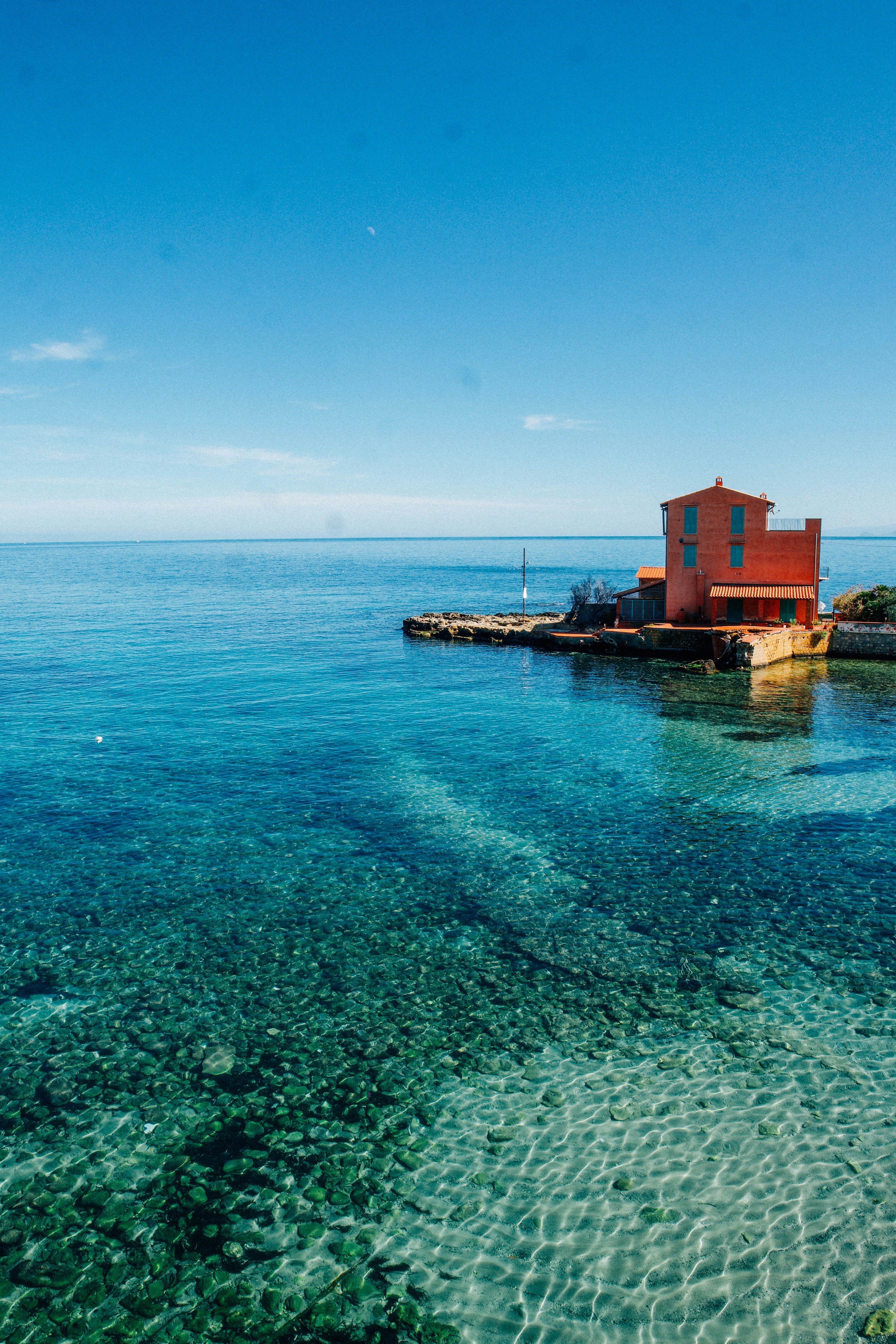 Red building on a rocky shore with clear blue water.