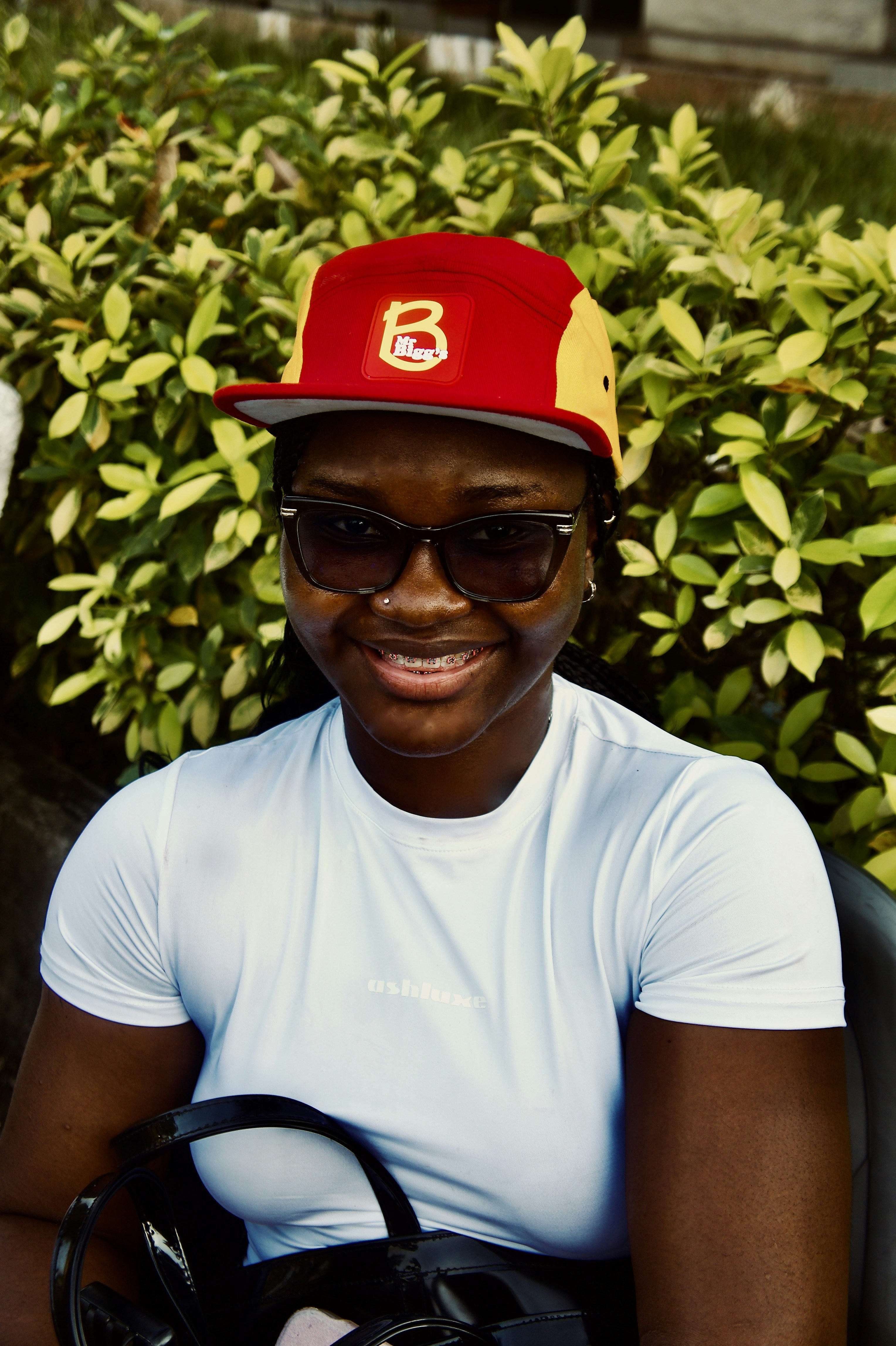 A woman wearing a red and yellow cap and glasses in front of green bushes