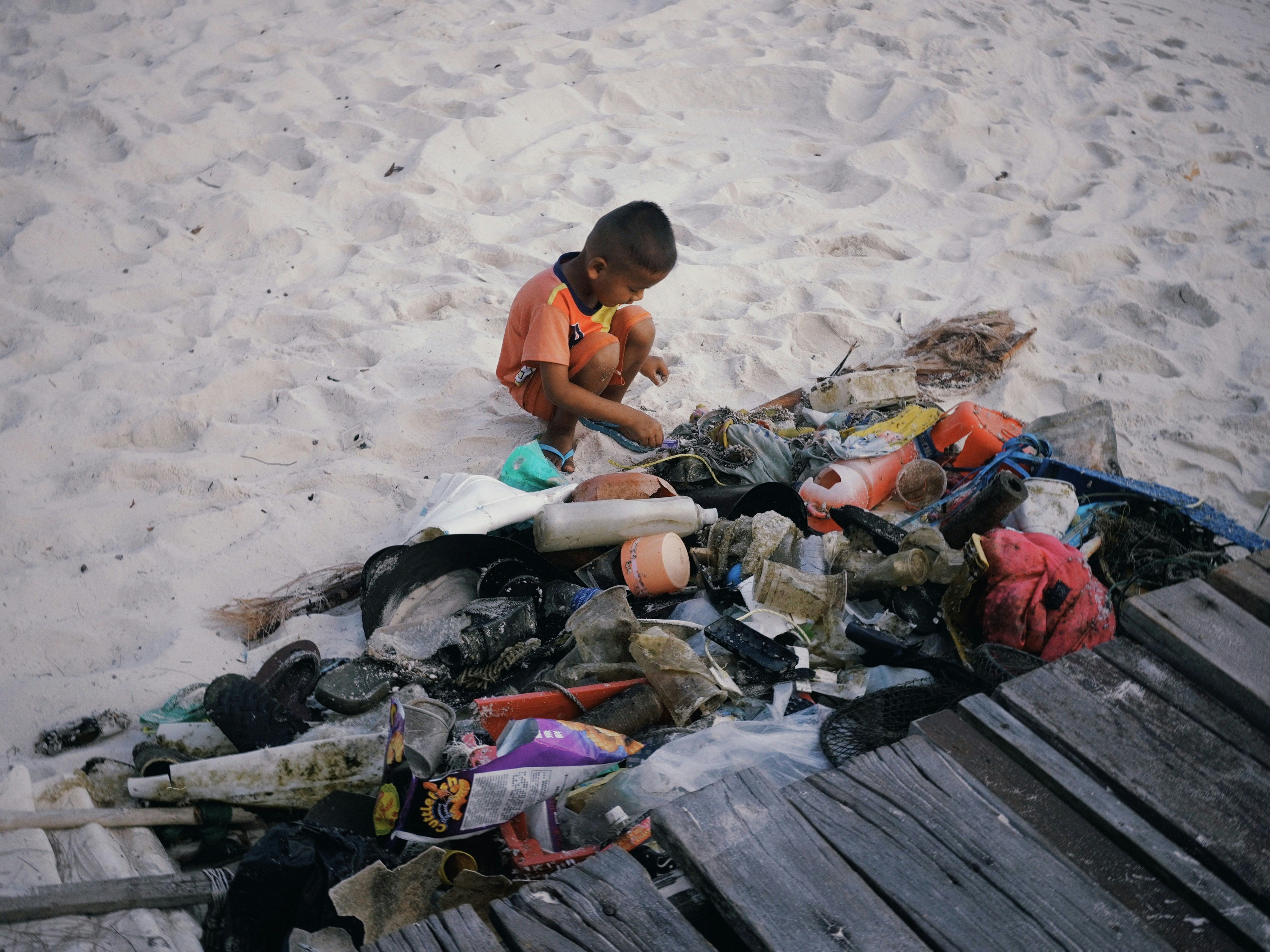 A young boy sits on a sandy beach with debris.