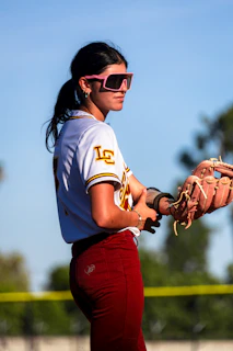 A young woman in softball uniform wearing pink sunglasses.