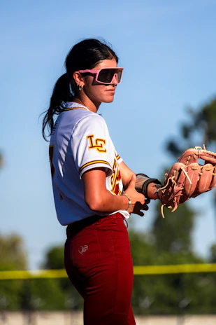 A young woman in softball uniform wearing pink sunglasses.