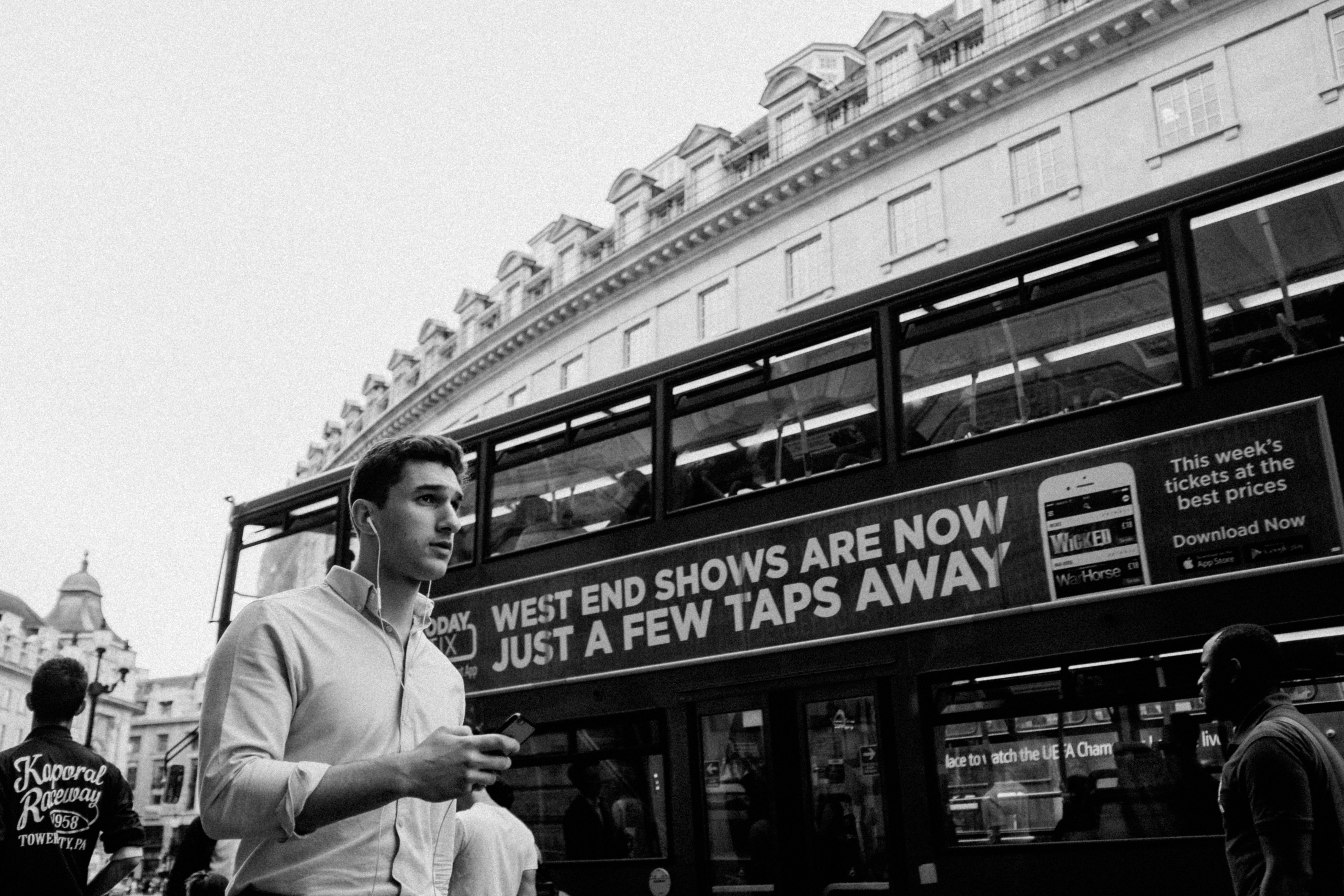 Man with headphones walks past double-decker bus in city.
