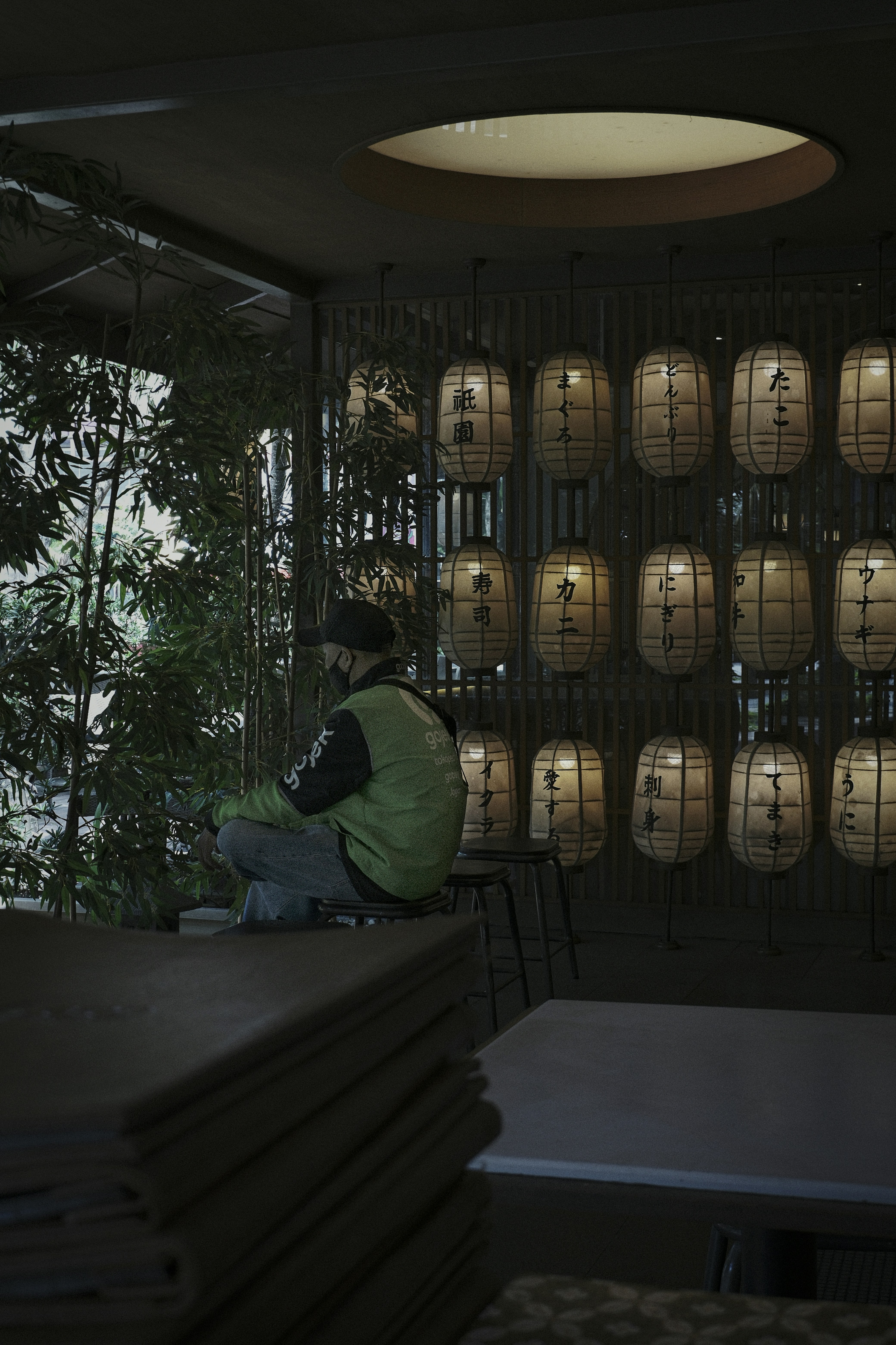 Man in mask sits near bamboo and lanterns.