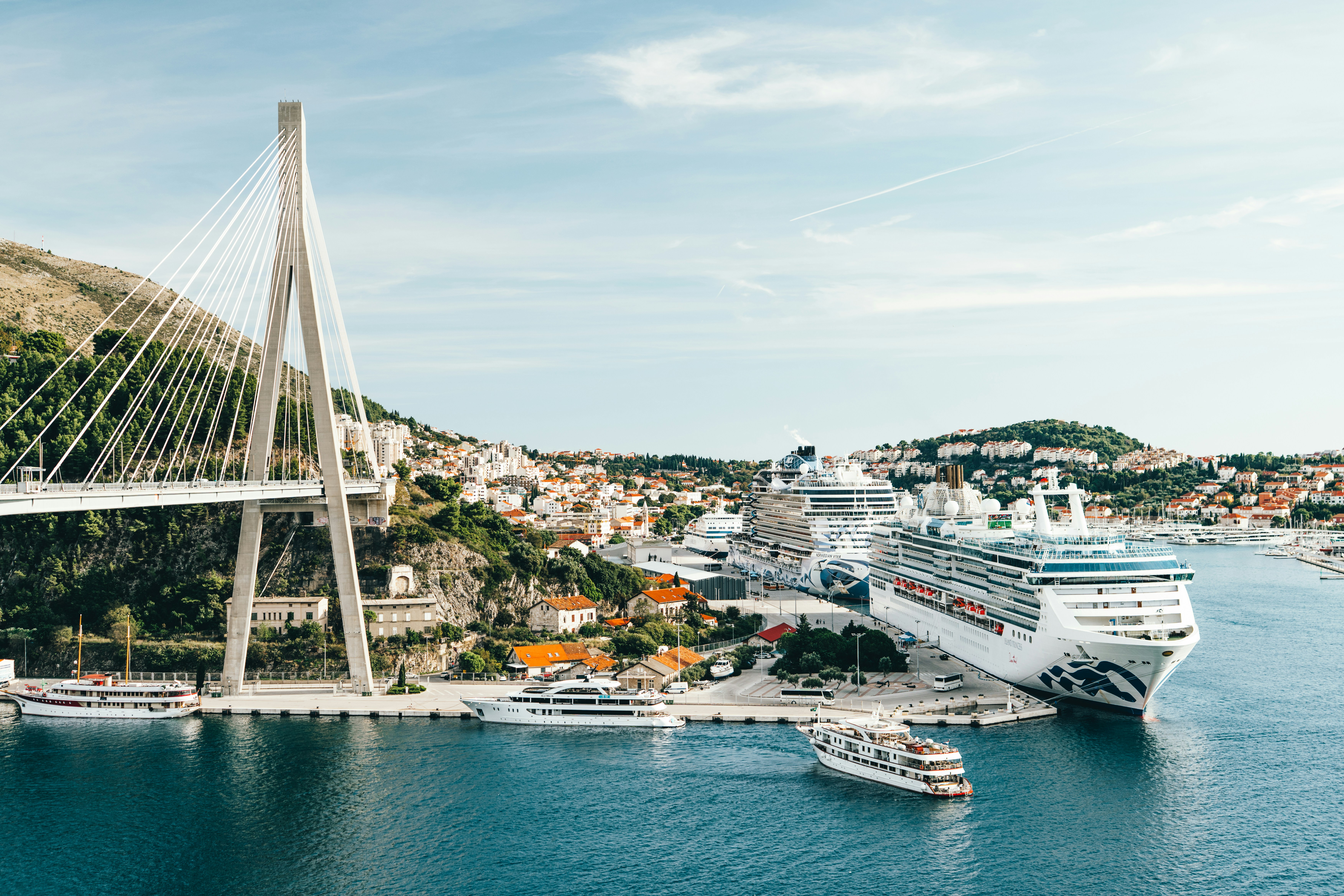 An aerial view of a large luxury cruise ship docked at a European port, with several smaller boats and emergency vehicles visible near the dock.