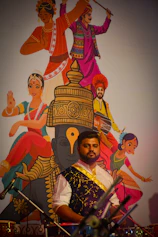 Man in traditional attire with cultural mural behind him