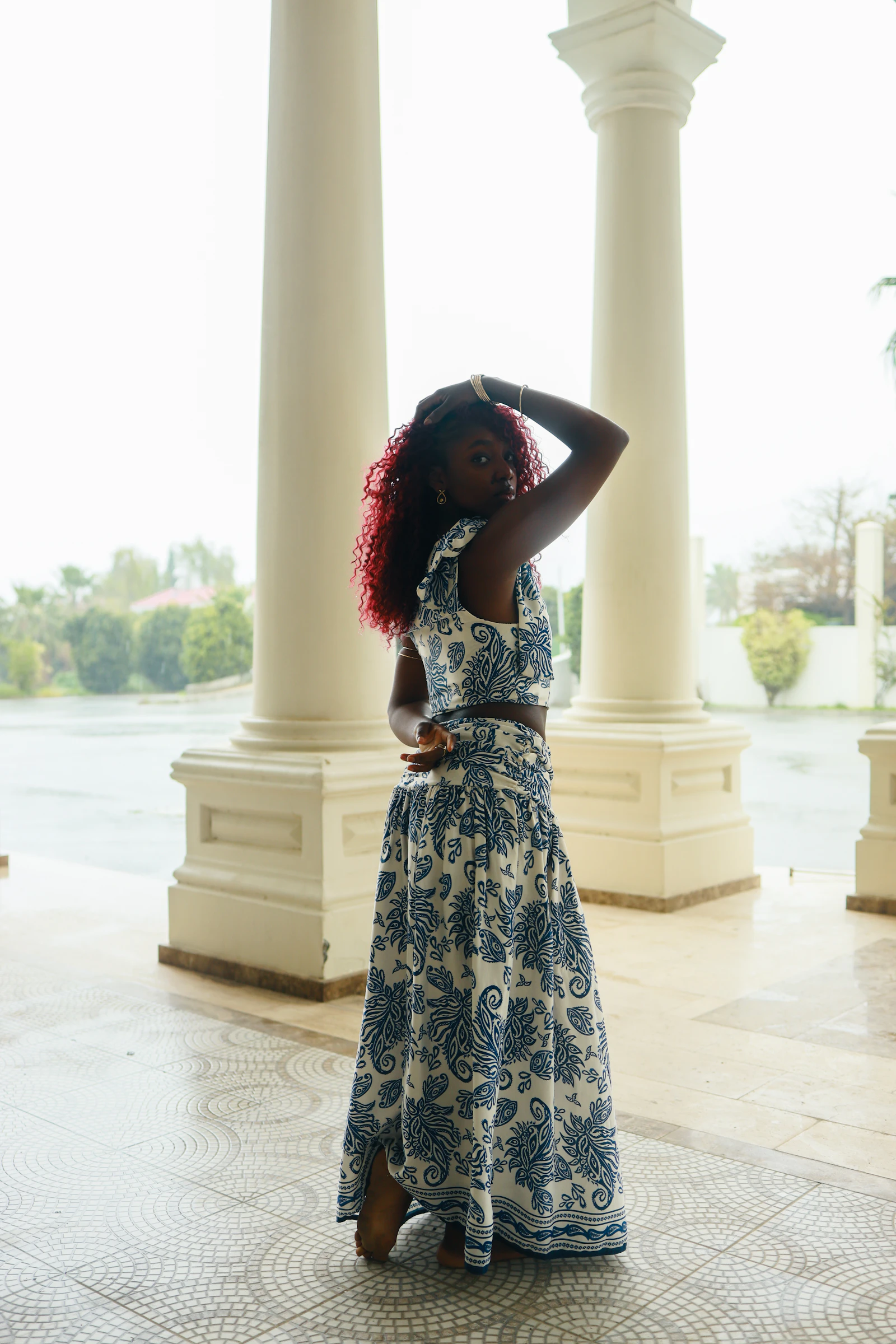 Editorial full-length portrait of a woman in a warm neutral two-piece outfit posing against sunlit columns — body-neutral, calm and abstract