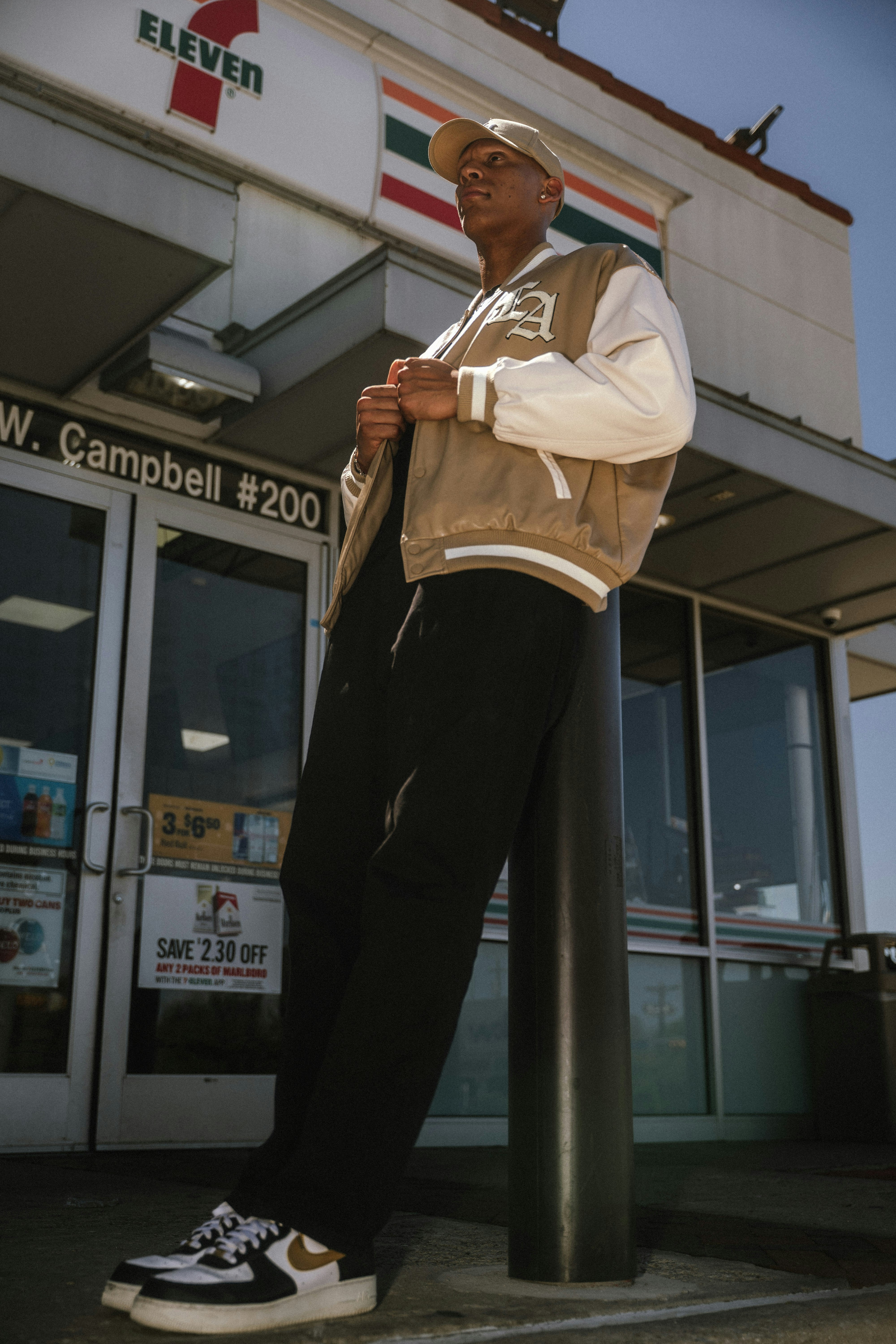 Man in varsity jacket leans against pole outside 7-eleven.