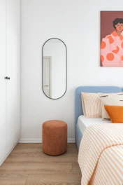 A minimalist bedroom with a blue bed and orange stool.