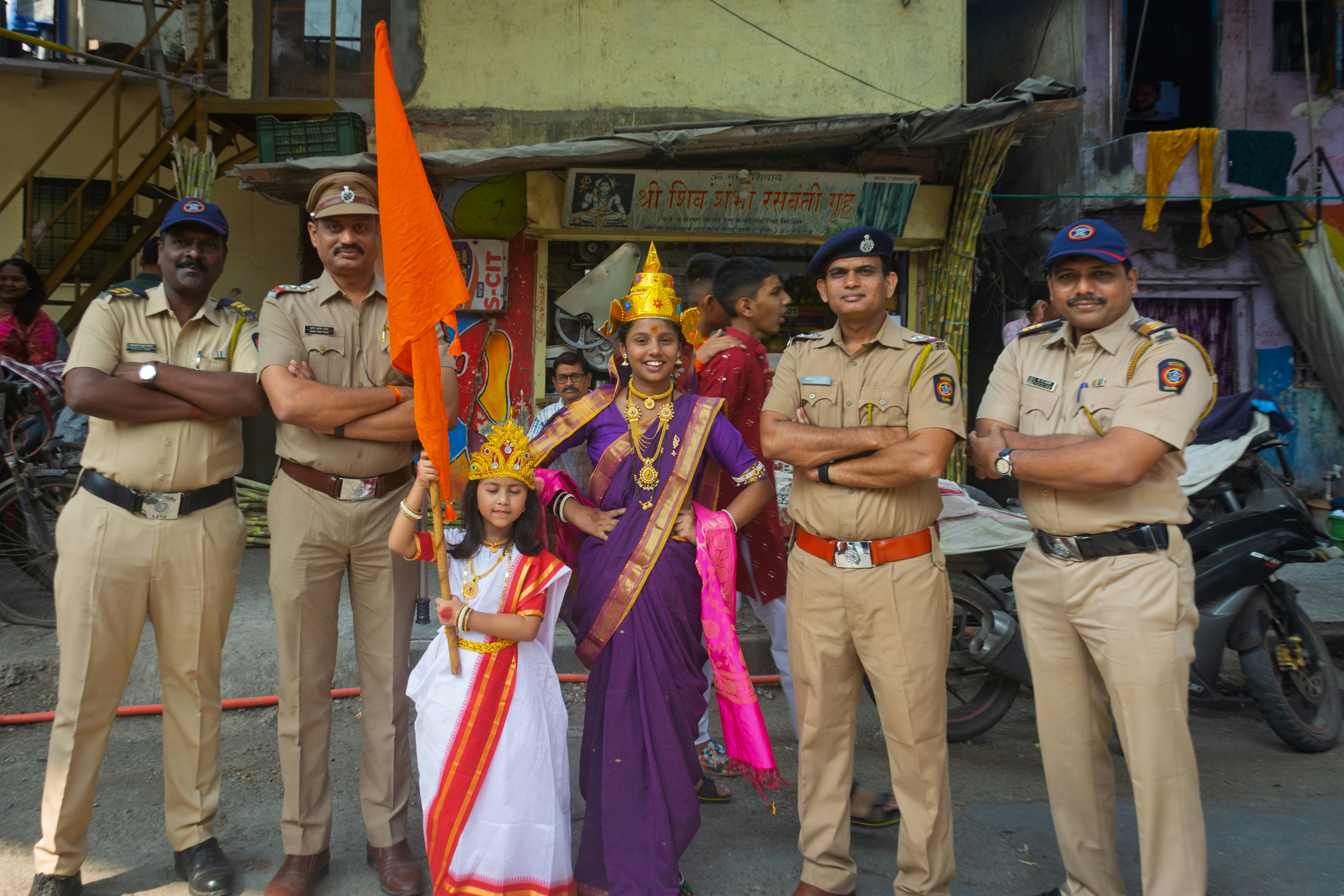 एक घर का दृश्य जो पुलिस टेप से घिरा हुआ है, आस-पास कुछ उत्सुक पड़ोसी और एक पुलिस अधिकारी किसी से बात करते हुए।