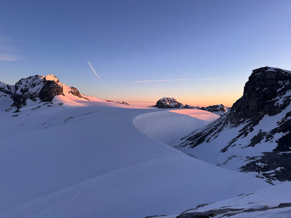 The image depicts a breathtaking view of a snow-covered mountain range at sunrise or sunset. The mountains, blanketed in pristine white snow, rise majestically against the sky, their peaks reaching towards the heavens. The sun is just beginning to set, casting a warm glow over the scene and painting the sky with hues of orange and pink. In the foreground, there are two small rocks that add a touch of ruggedness to the otherwise smooth landscape.