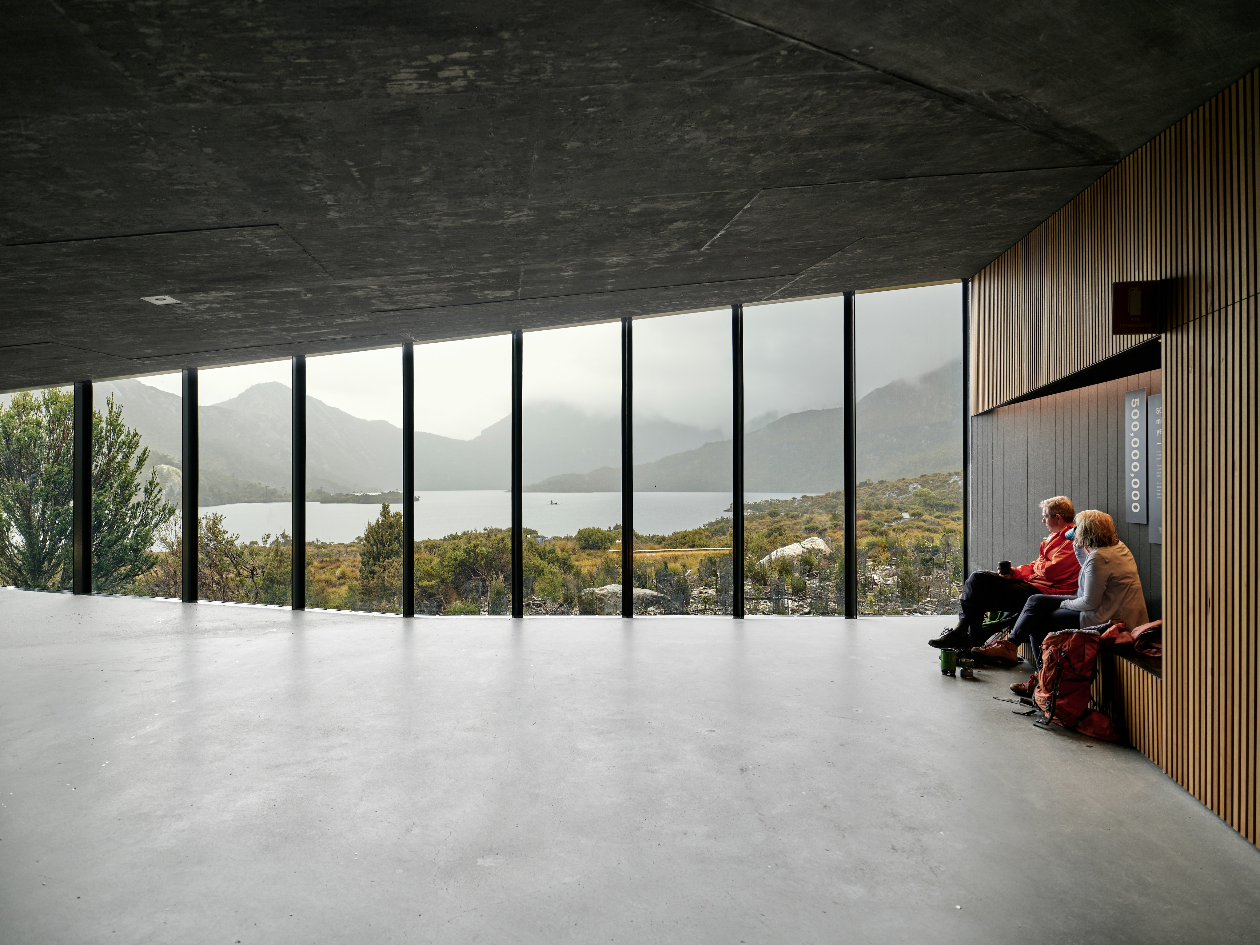 Two people relax inside a modern building overlooking a lake.