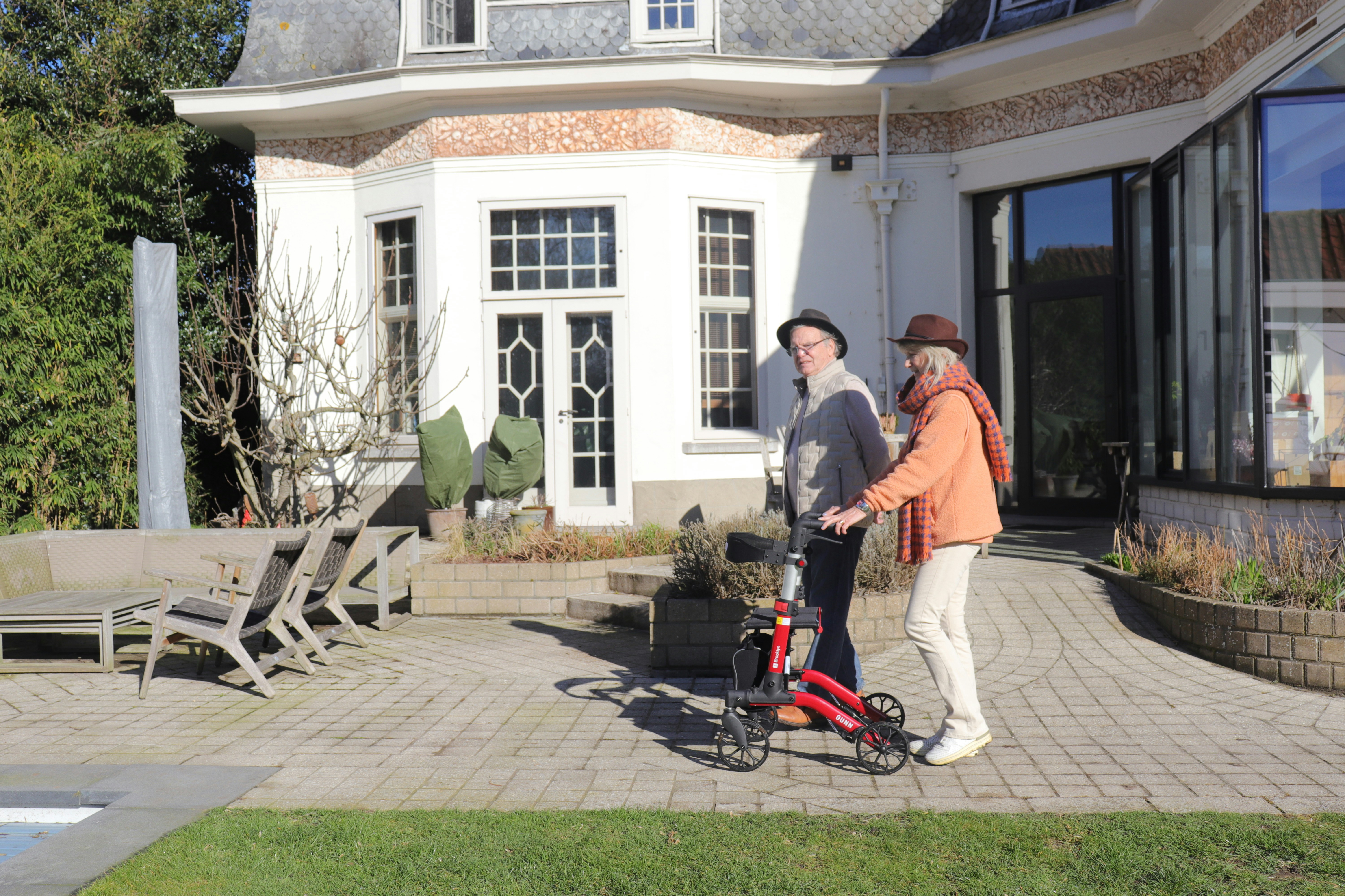 Two elderly people walk with a red rollator on a sunny paved patio next to a large elegant house and garden.