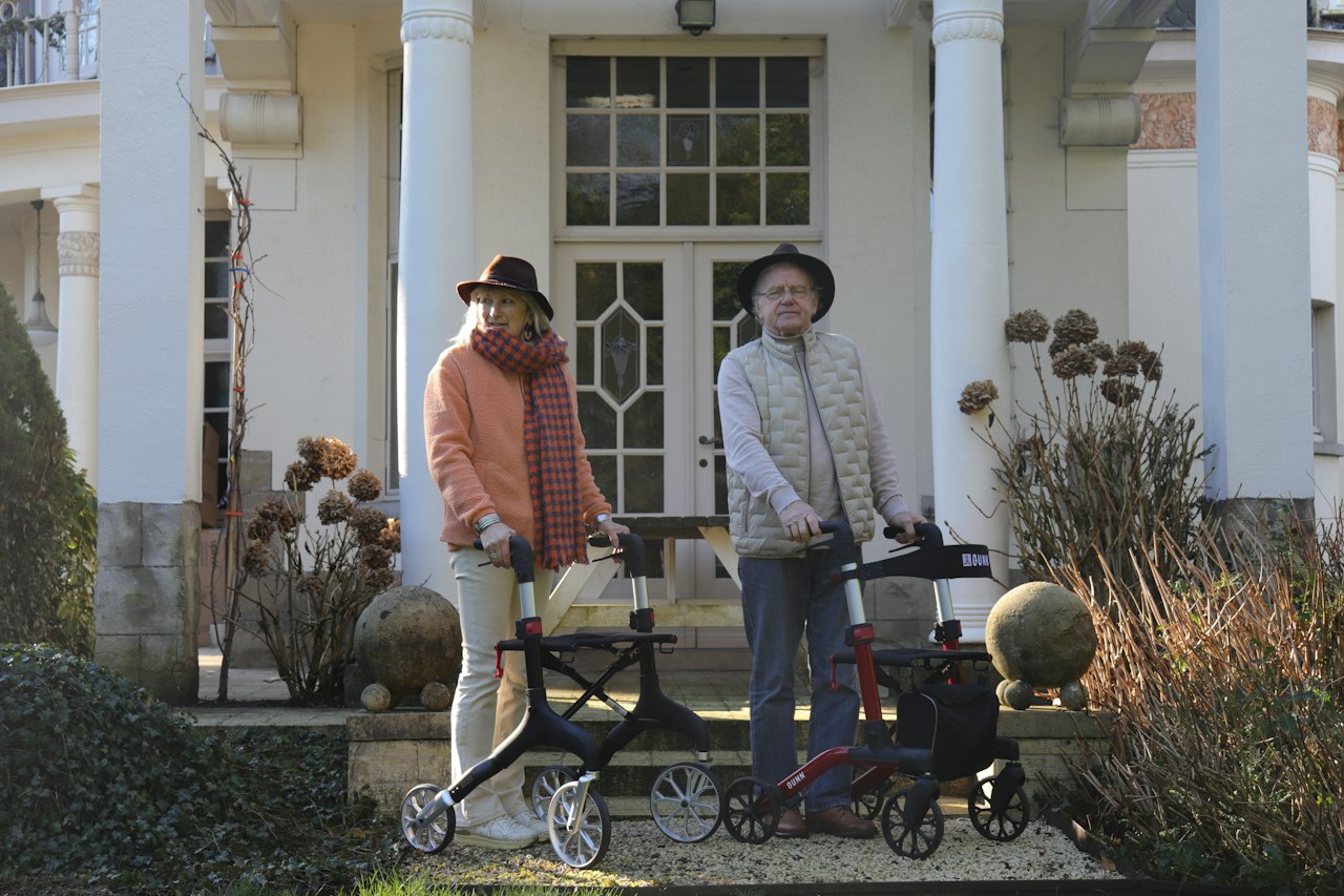 Two older adults with rollator walkers outside a columned house