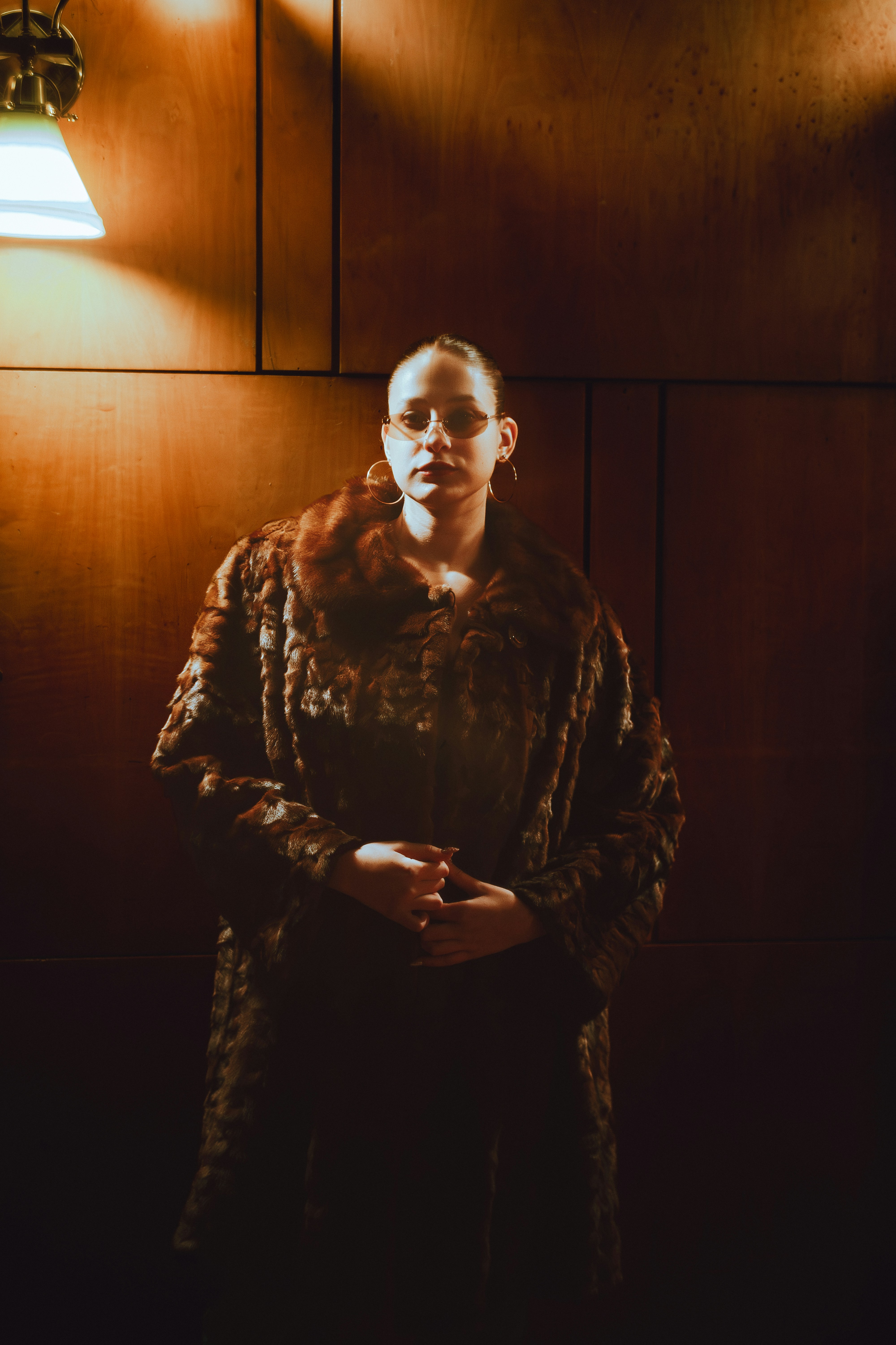 Woman in fur coat stands against wood-paneled wall