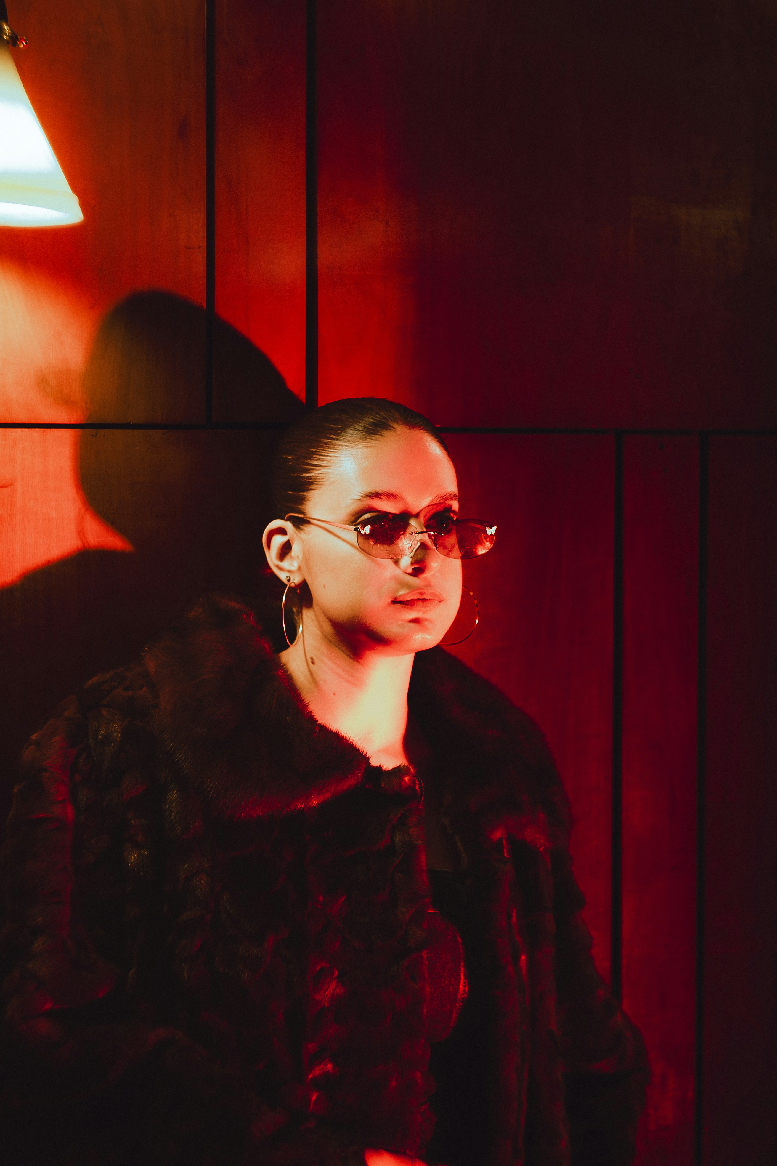 Woman in sunglasses and fur coat under red light.