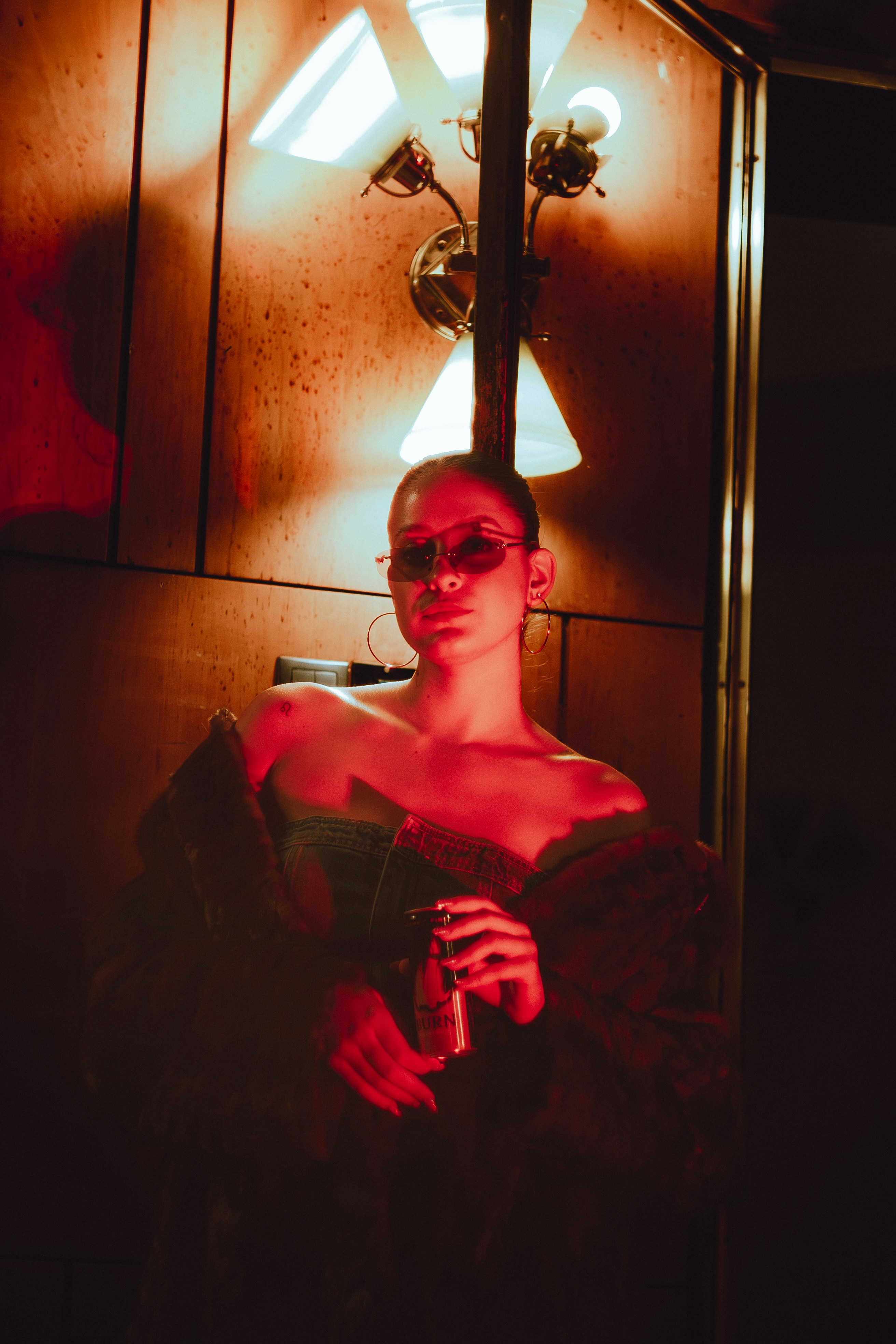 Woman in sunglasses holding a drink under red lights.