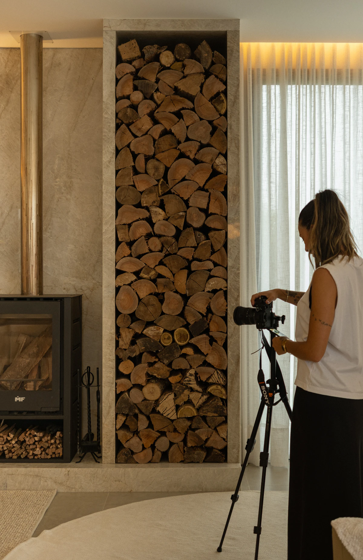 A woman sets up a camera on a tripod beside a stacked-log fireplace and sheer curtains in a calm living room