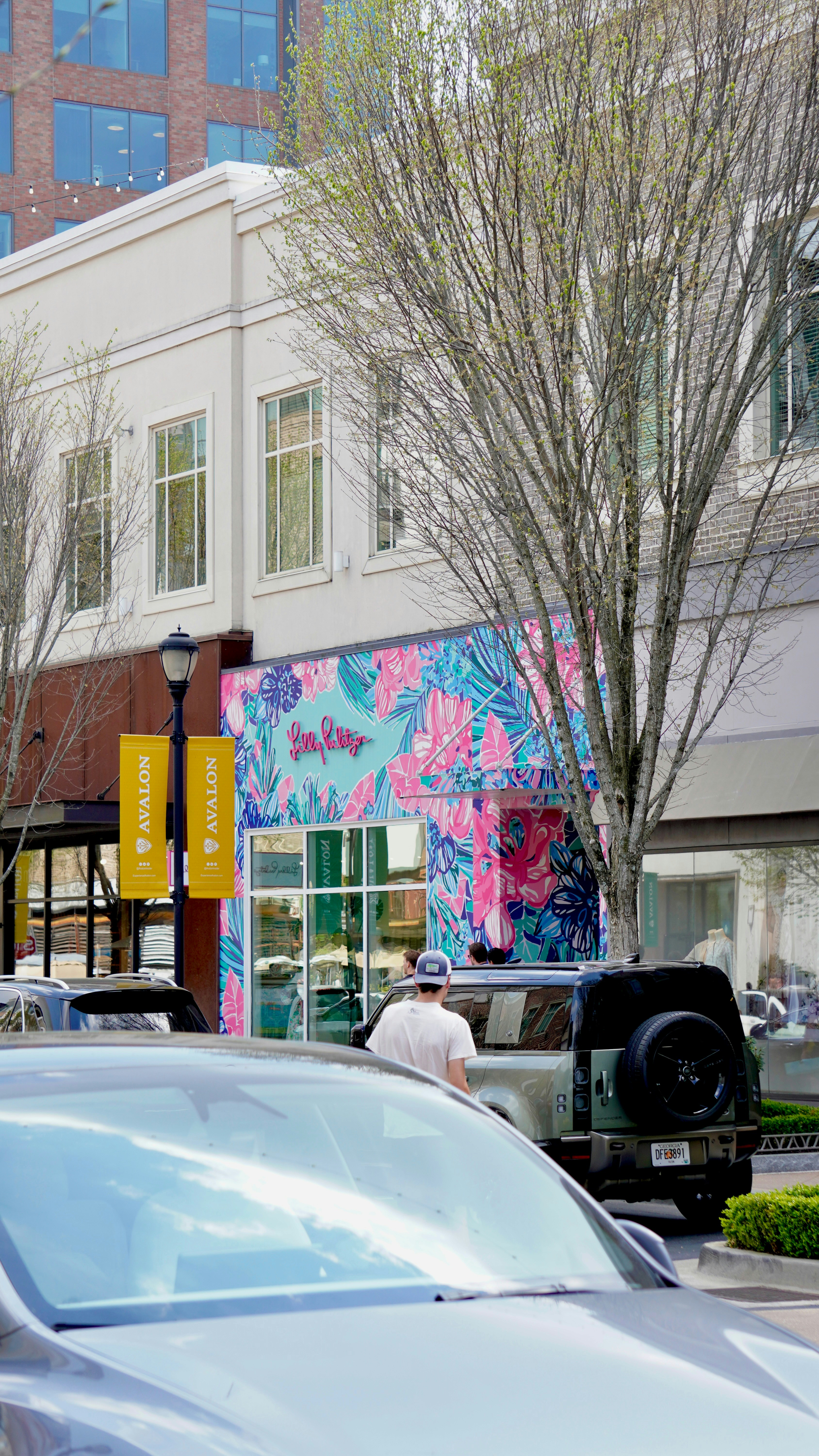 A man stands near a colorful storefront with cars parked.