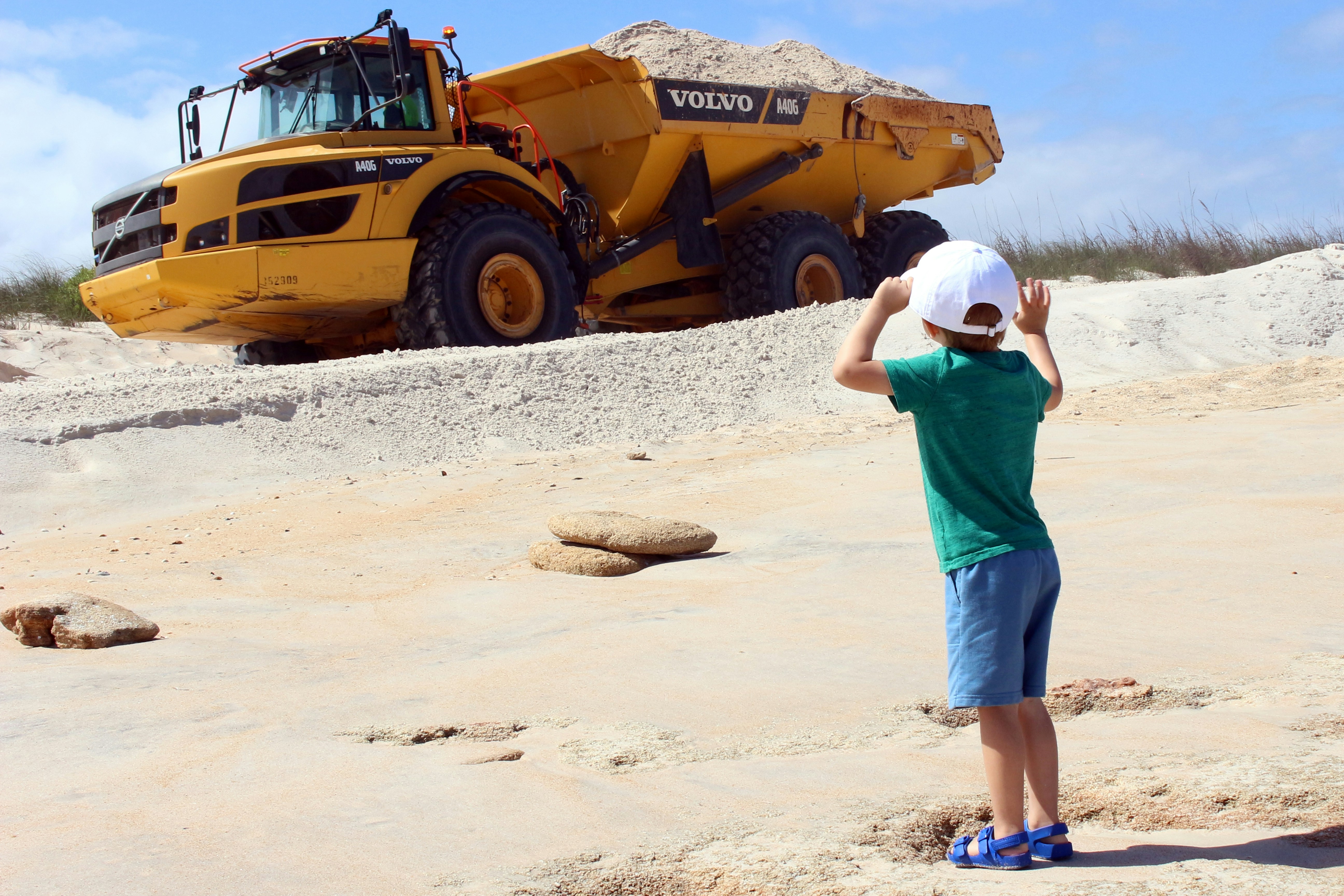 A child watches a large yellow dump truck in sand.