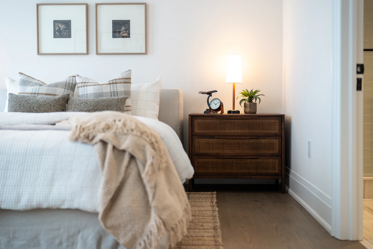 Cozy apartment bedroom with warm nightstand lamp glow, plaid throw pillows, woven cane dresser and framed wall art