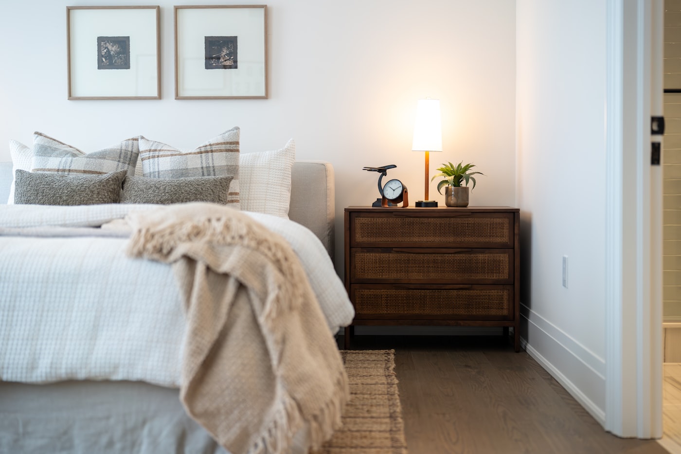 Warm beige apartment bedroom with layered plaid and woven linen pillows, soft cream throw blanket, rattan-front nightstand, and glowing bedside lamp on natural wood floors