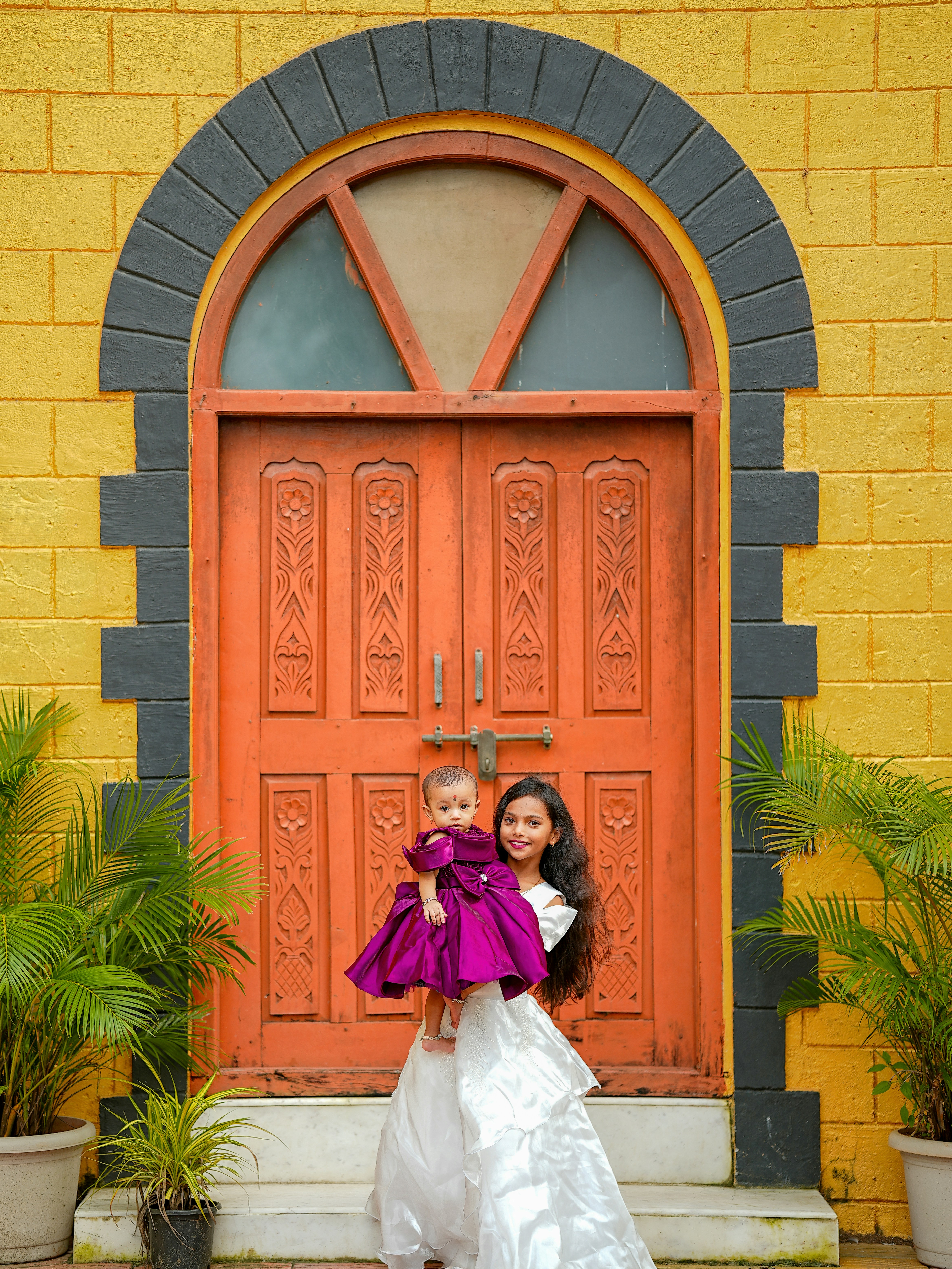 Two sisters in dresses stand before an orange door.