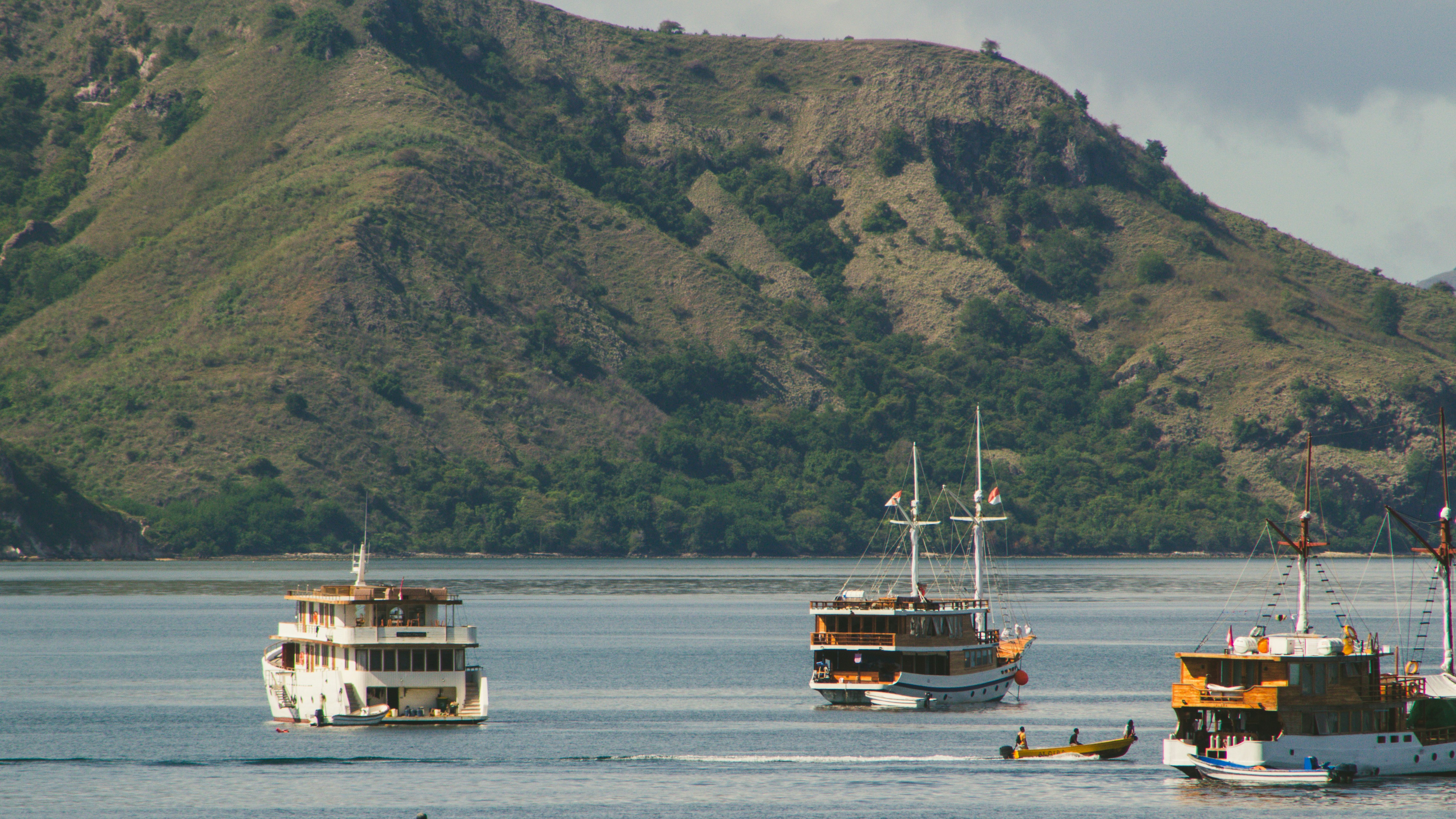 Several boats float on the calm ocean near hills.