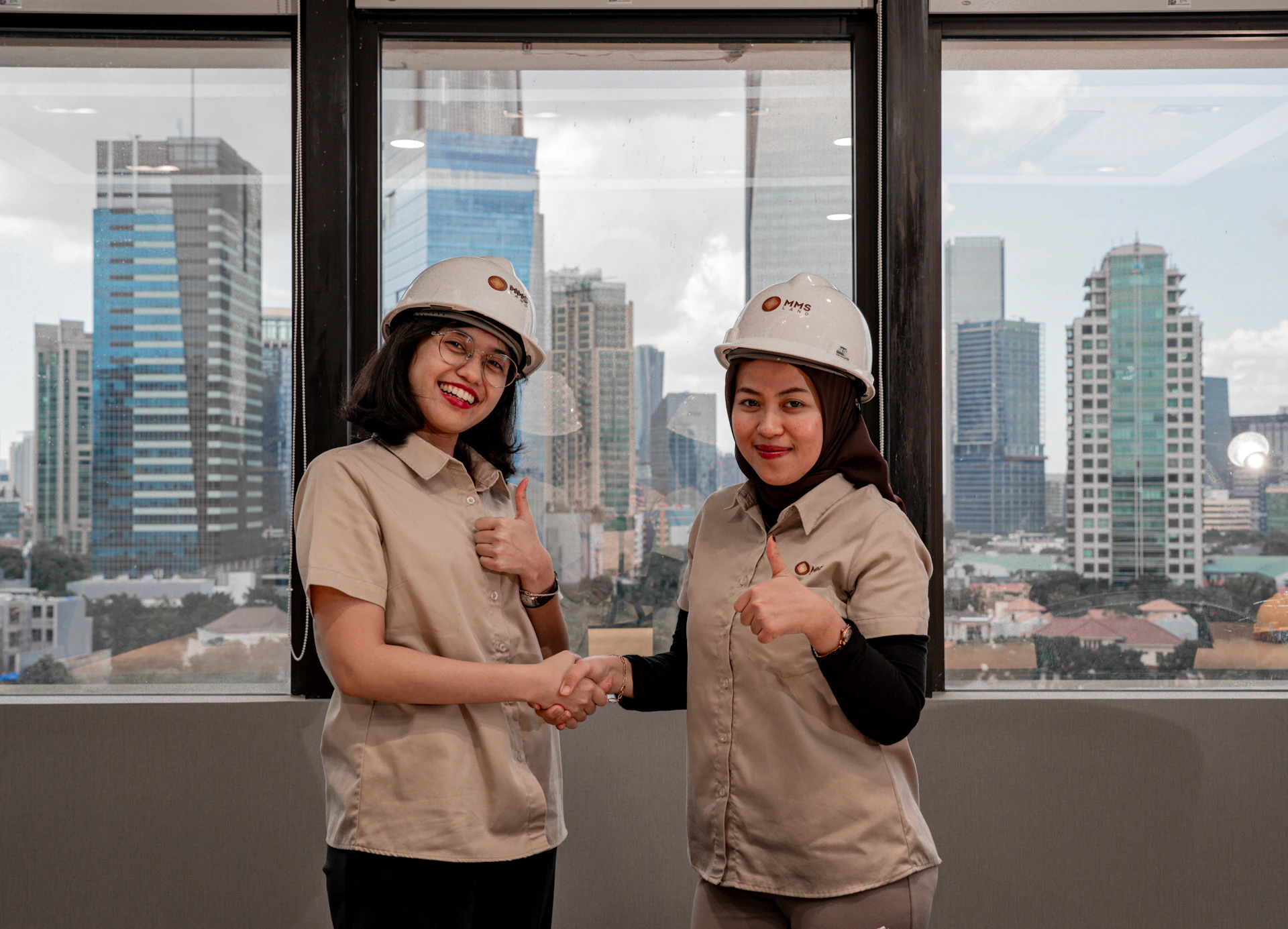 Two women in hard hats shake hands with city skyline.