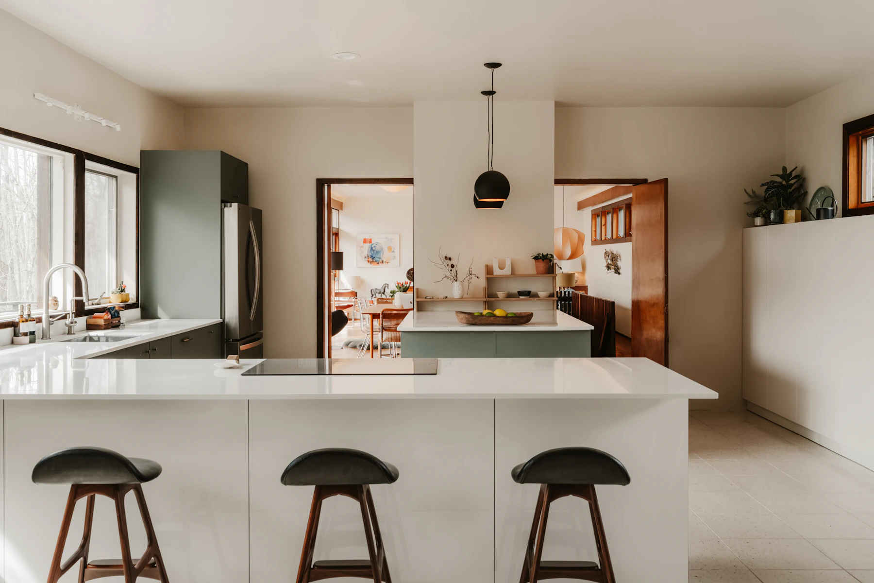 Kitchen with pale counters, muted green cabinetry, black pendants and stools with wooden legs