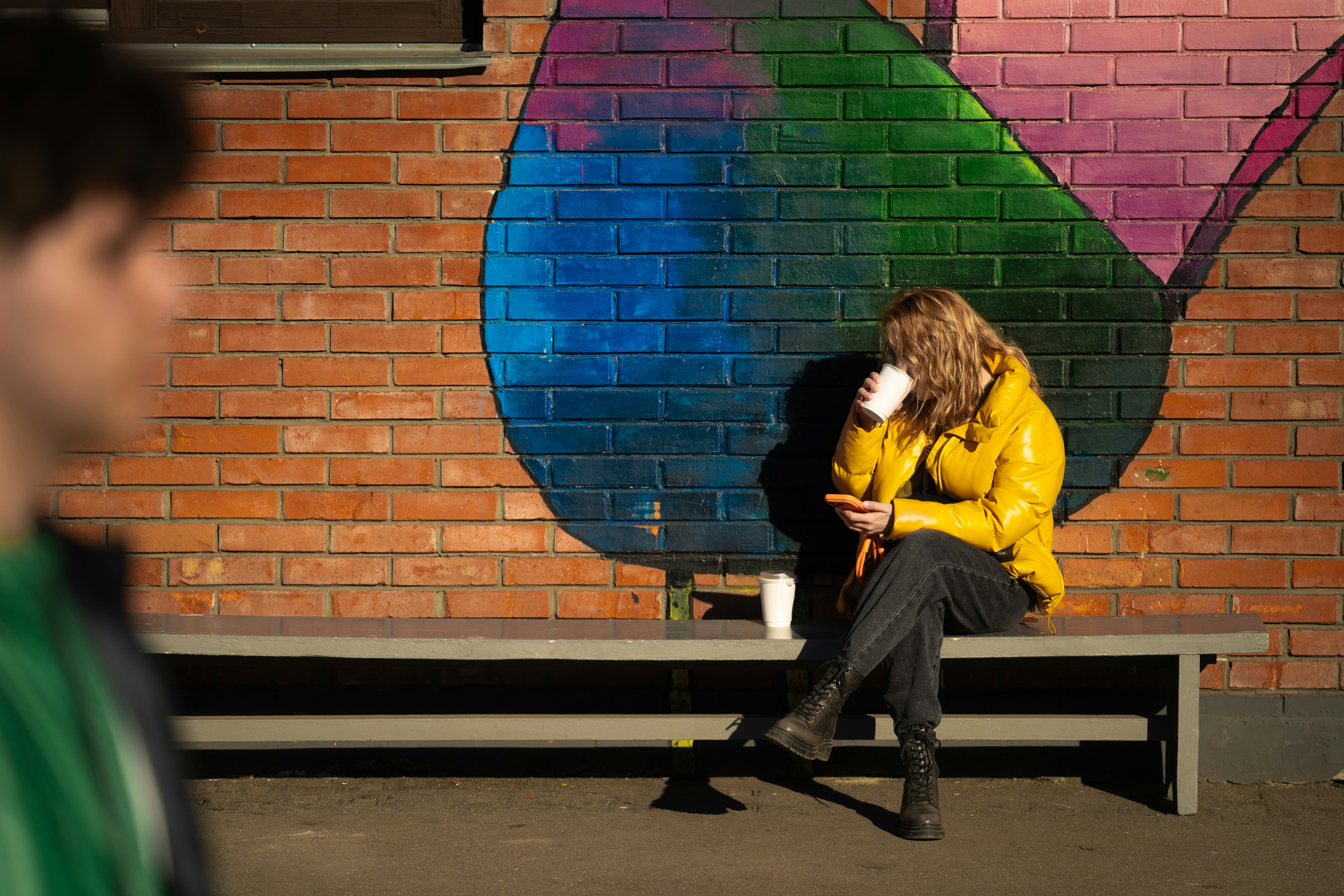 Frau in gelber Jacke sitzt auf einer Bank in der Nähe des Wandgemäldes
