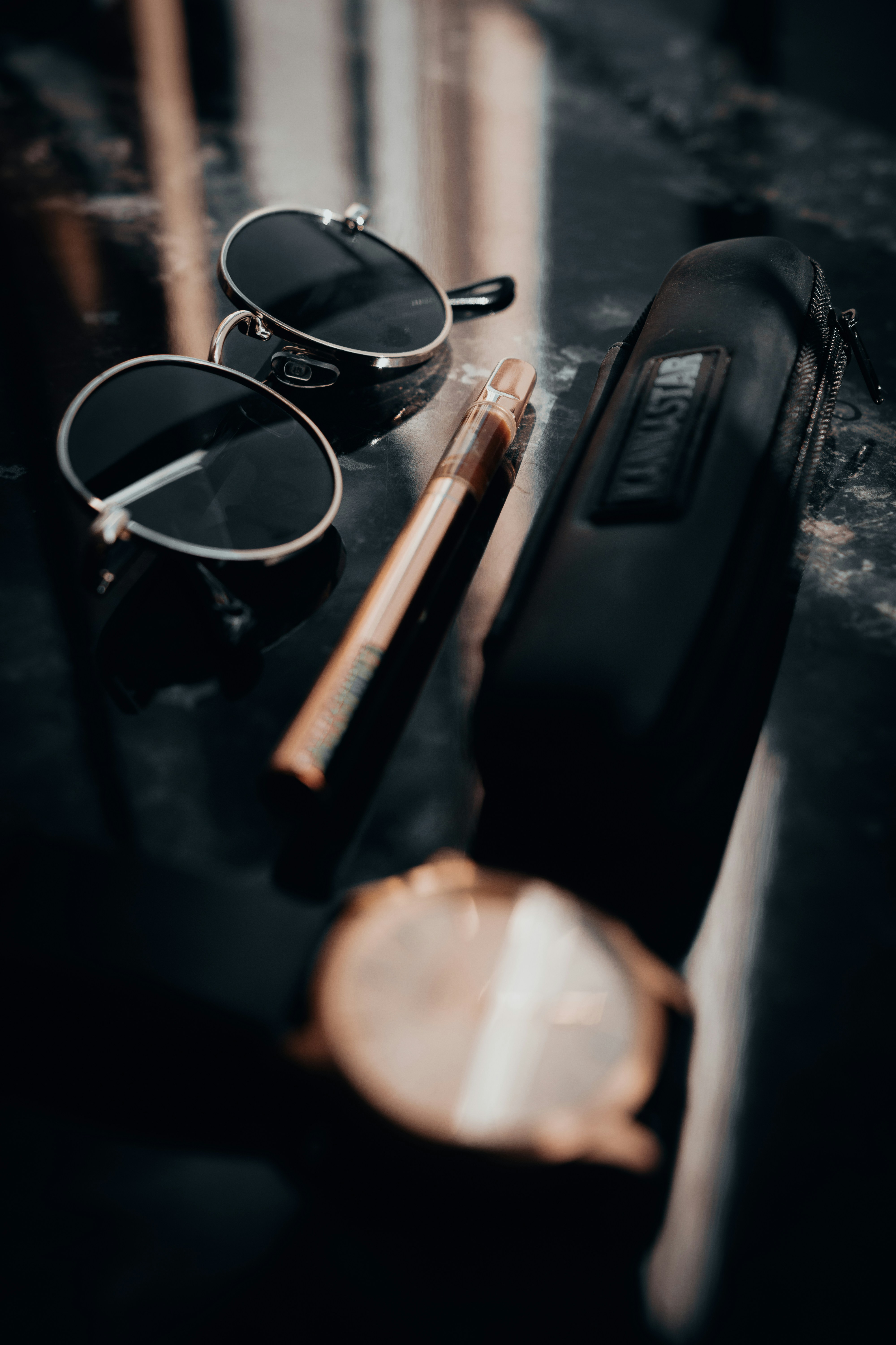 Round sunglasses, vape pen, and watch on dark surface