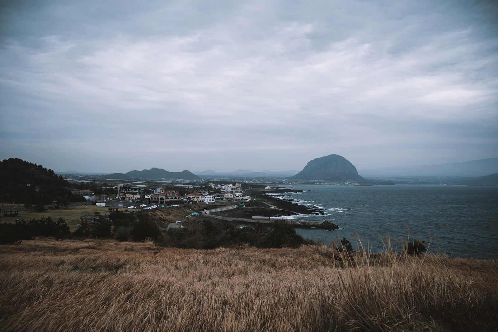 A Jeju coastal town below an overcast sky and distant volcanic peak