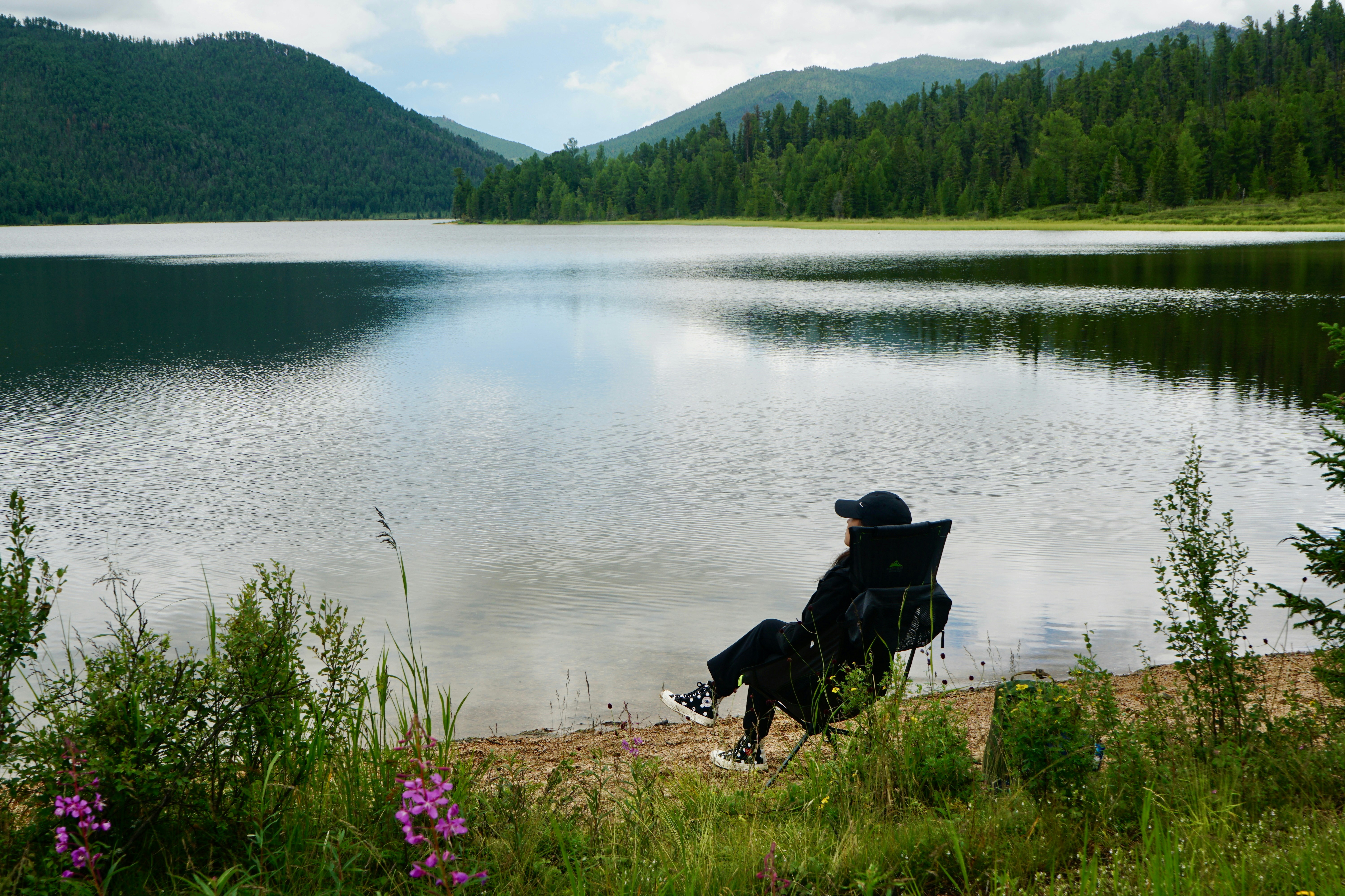Person relaxing by a serene lake with forested mountains.