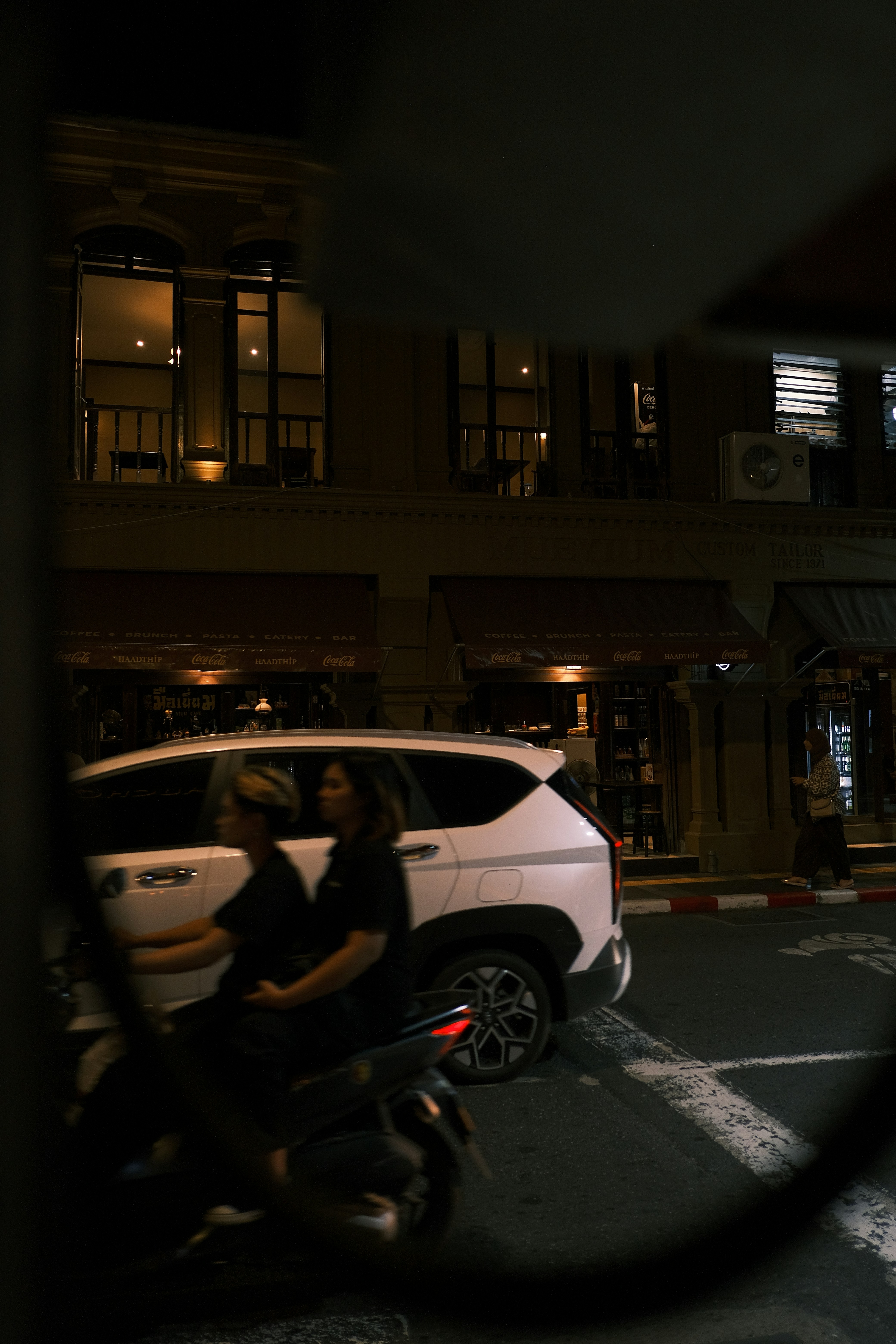 Two people ride a scooter at night past buildings.