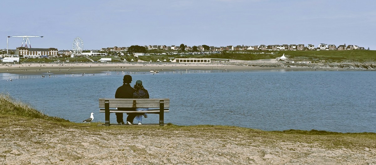 Couple sitting on a bench by the beach, overlooking the water and town, discussing beach cleanup tips and ideas.