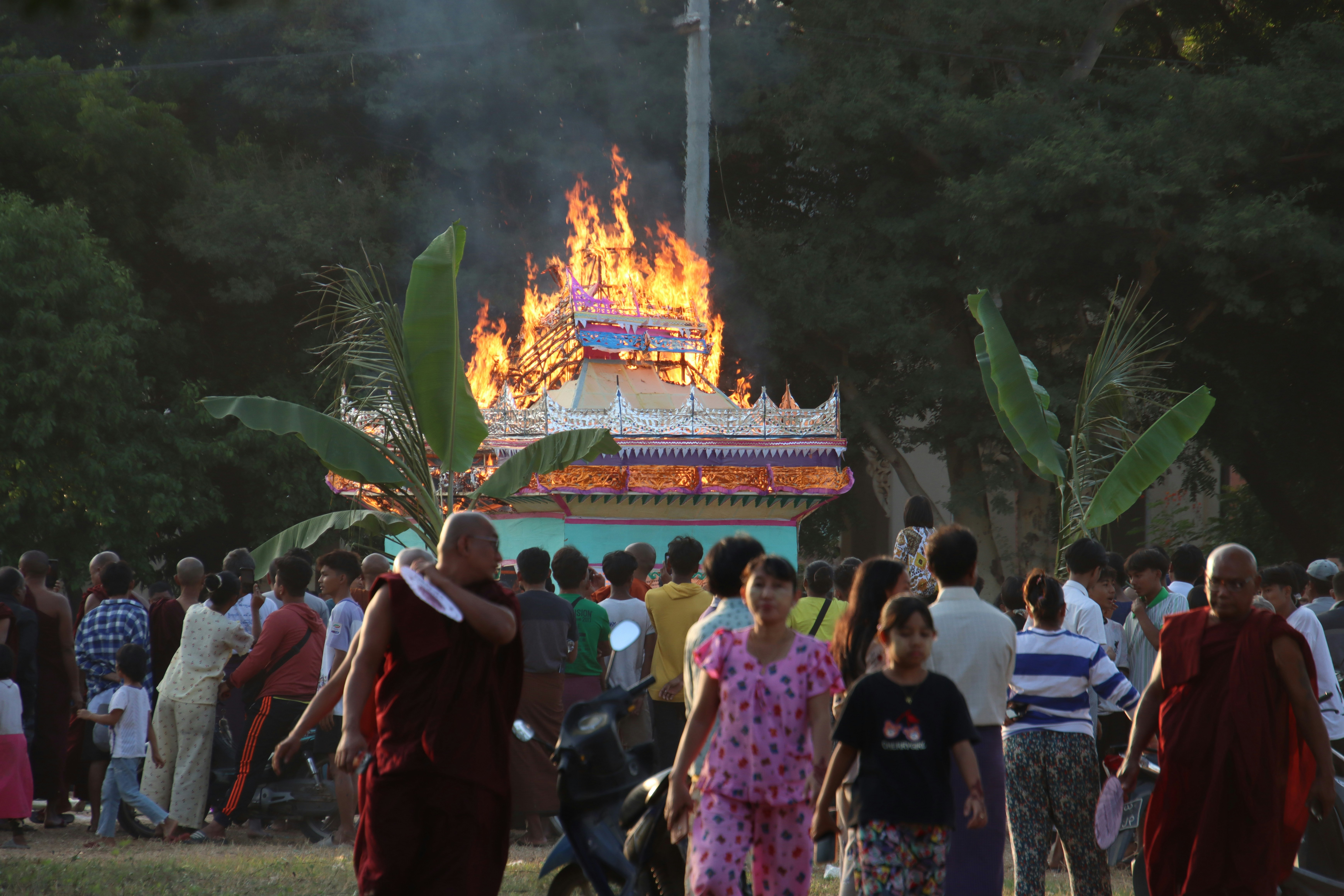 A funeral pyre burns intensely with a crowd watching
