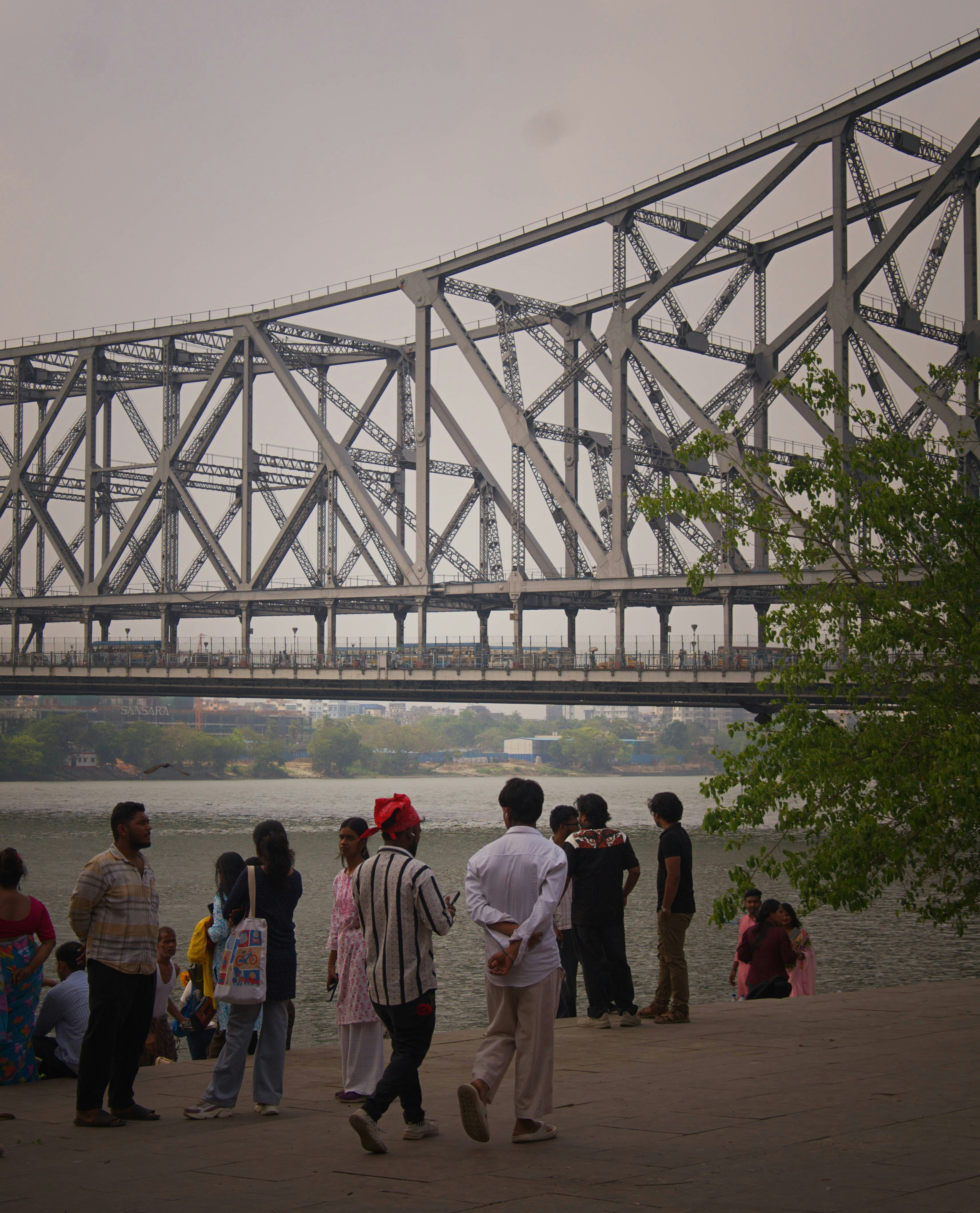 People gather near a large metal bridge over a river.