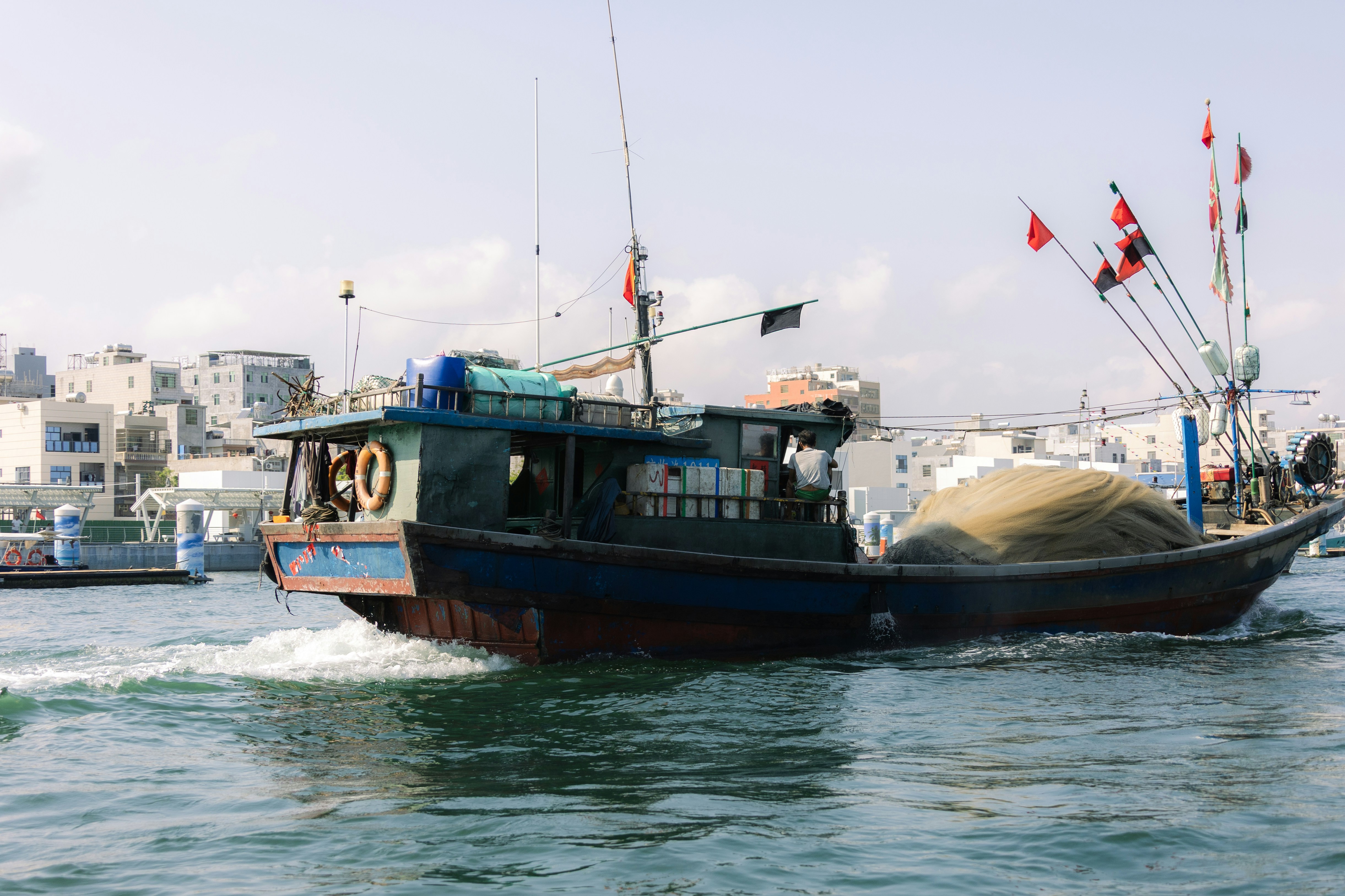 A fishing boat sails on the water with buildings behind.