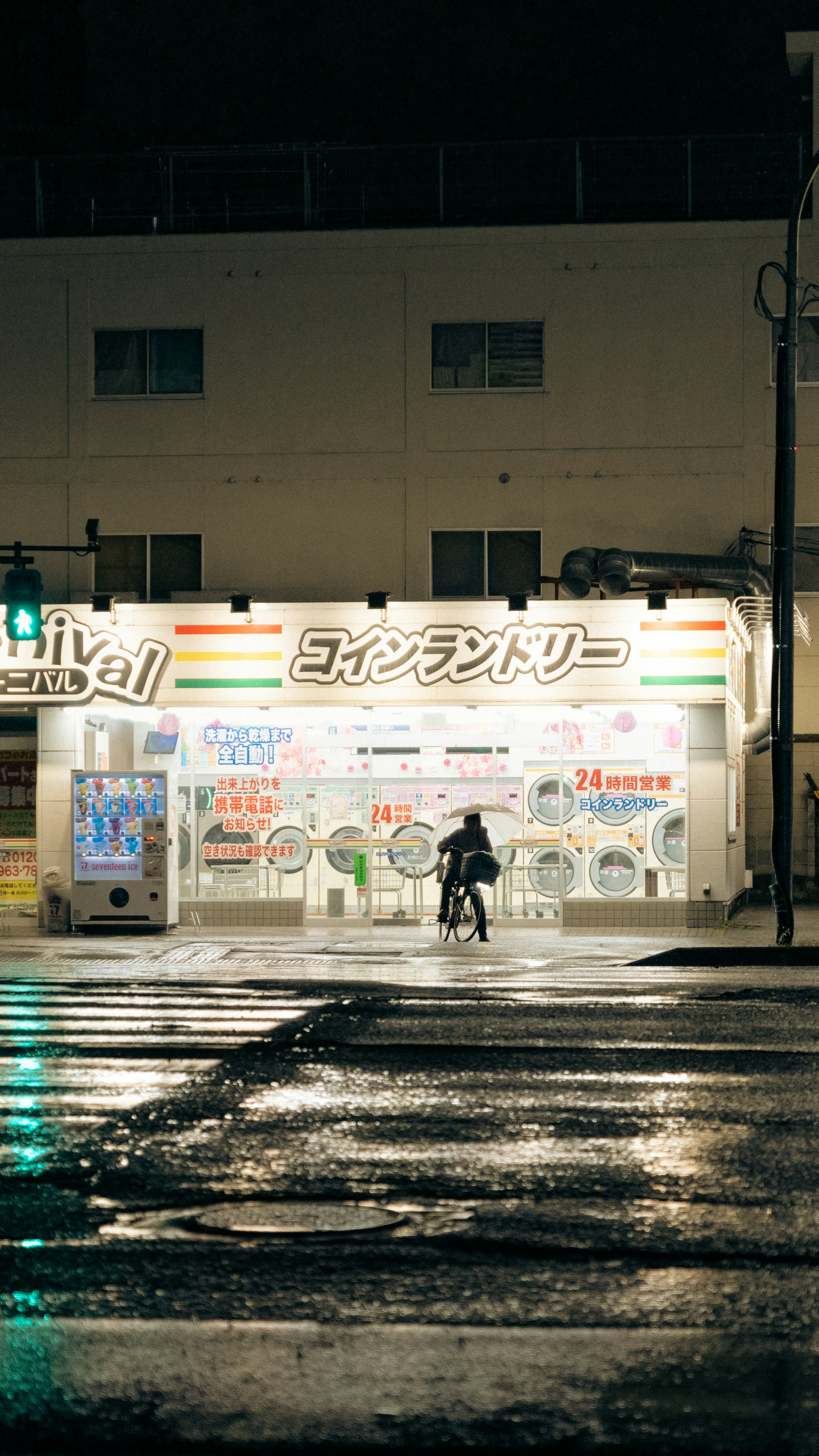 Person rides bicycle past brightly lit coin laundry at night.