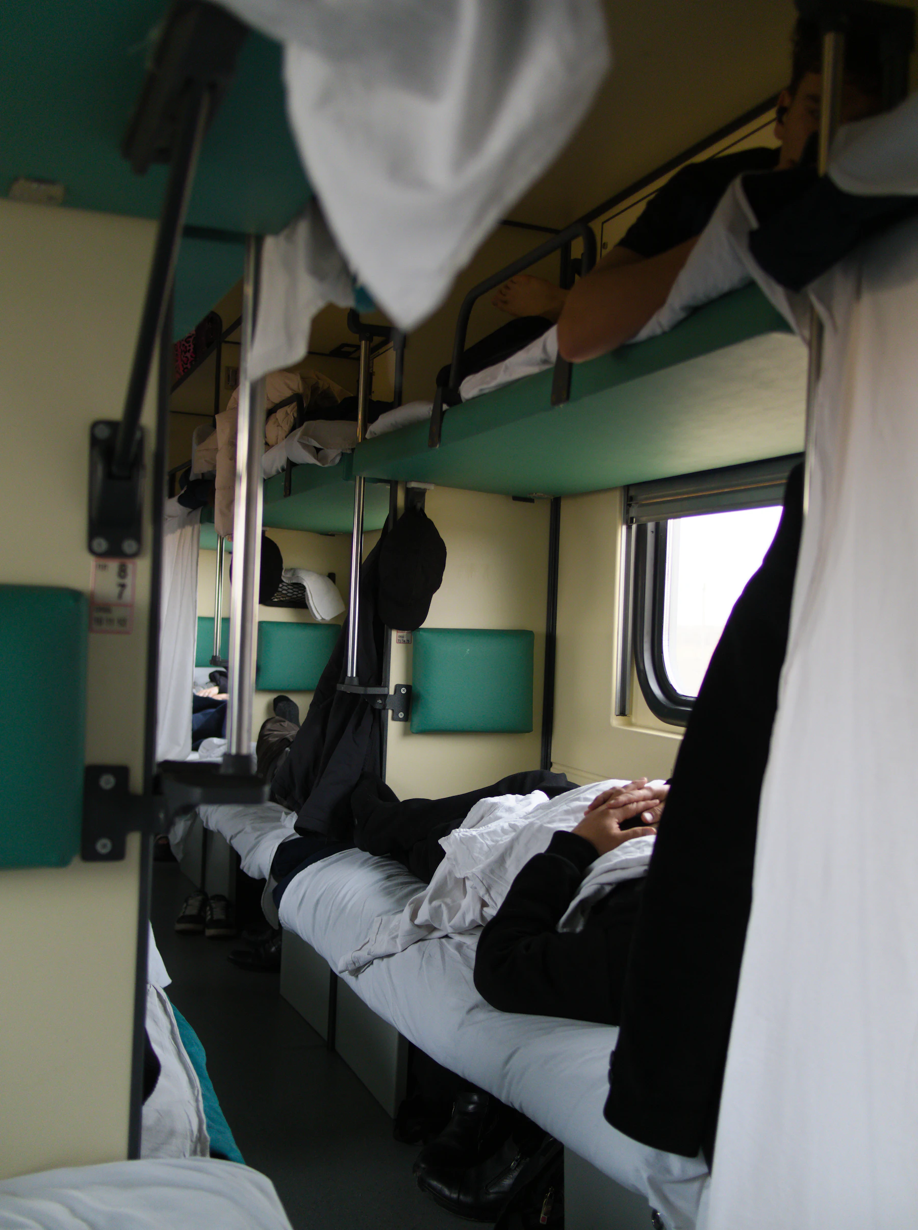 Inside a sleeper train car, passengers lying on green bunk berths that line the aisle