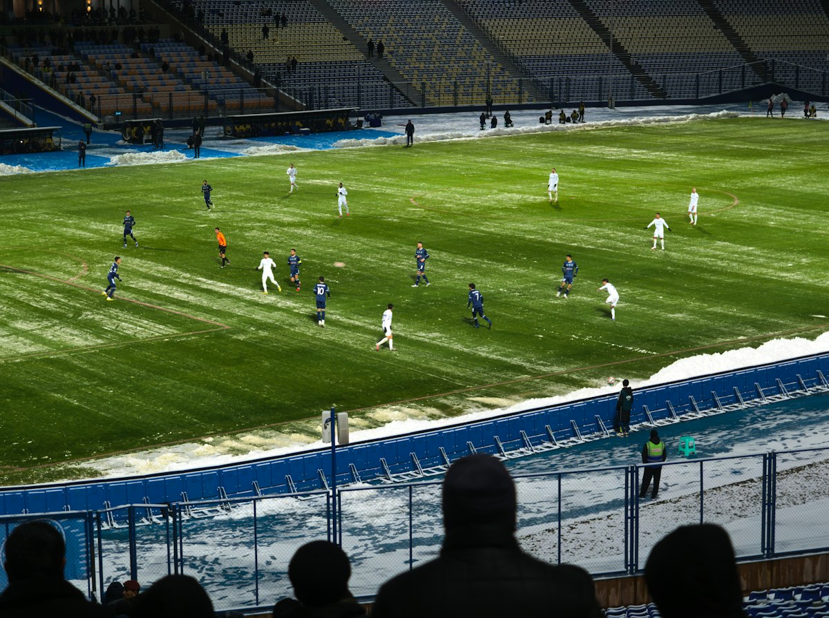 Match de football joué sur une pelouse bordée de neige dans un stade, vu depuis les tribunes derrière des spectateurs