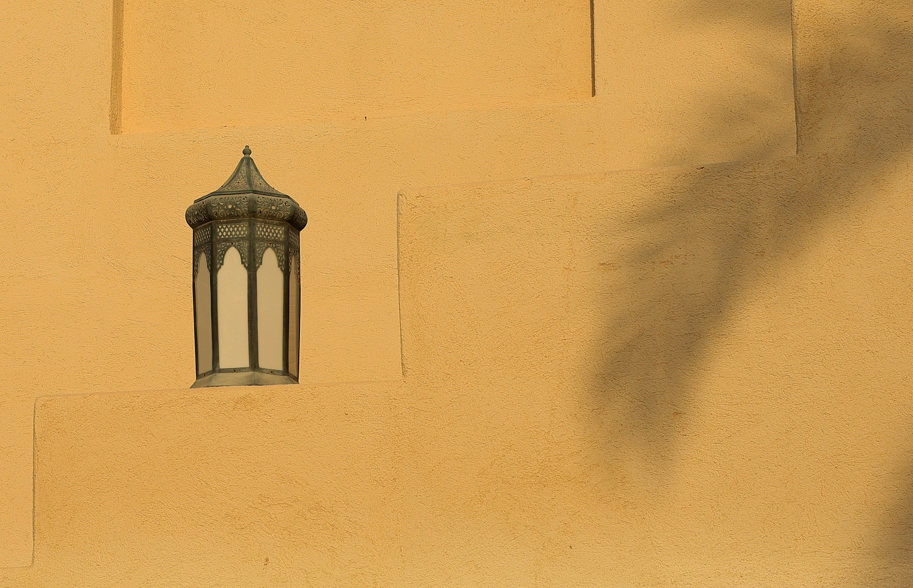 Ornate lantern on a textured yellow wall with palm shadow.