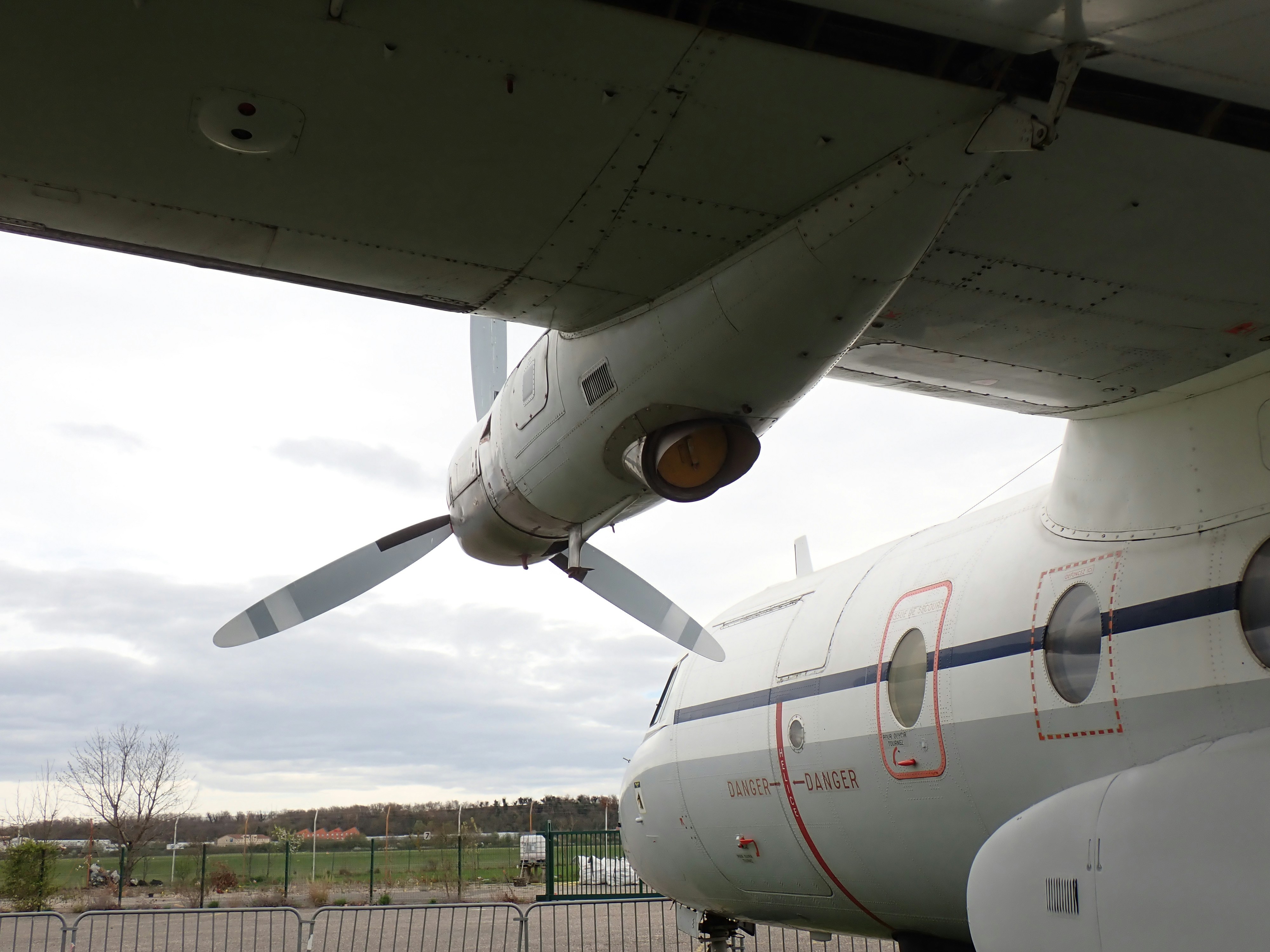Under-wing view of a twin-engine regional aircraft, showcasing the engine nacelle and fuselage markings with a distant runway and fence in the background.